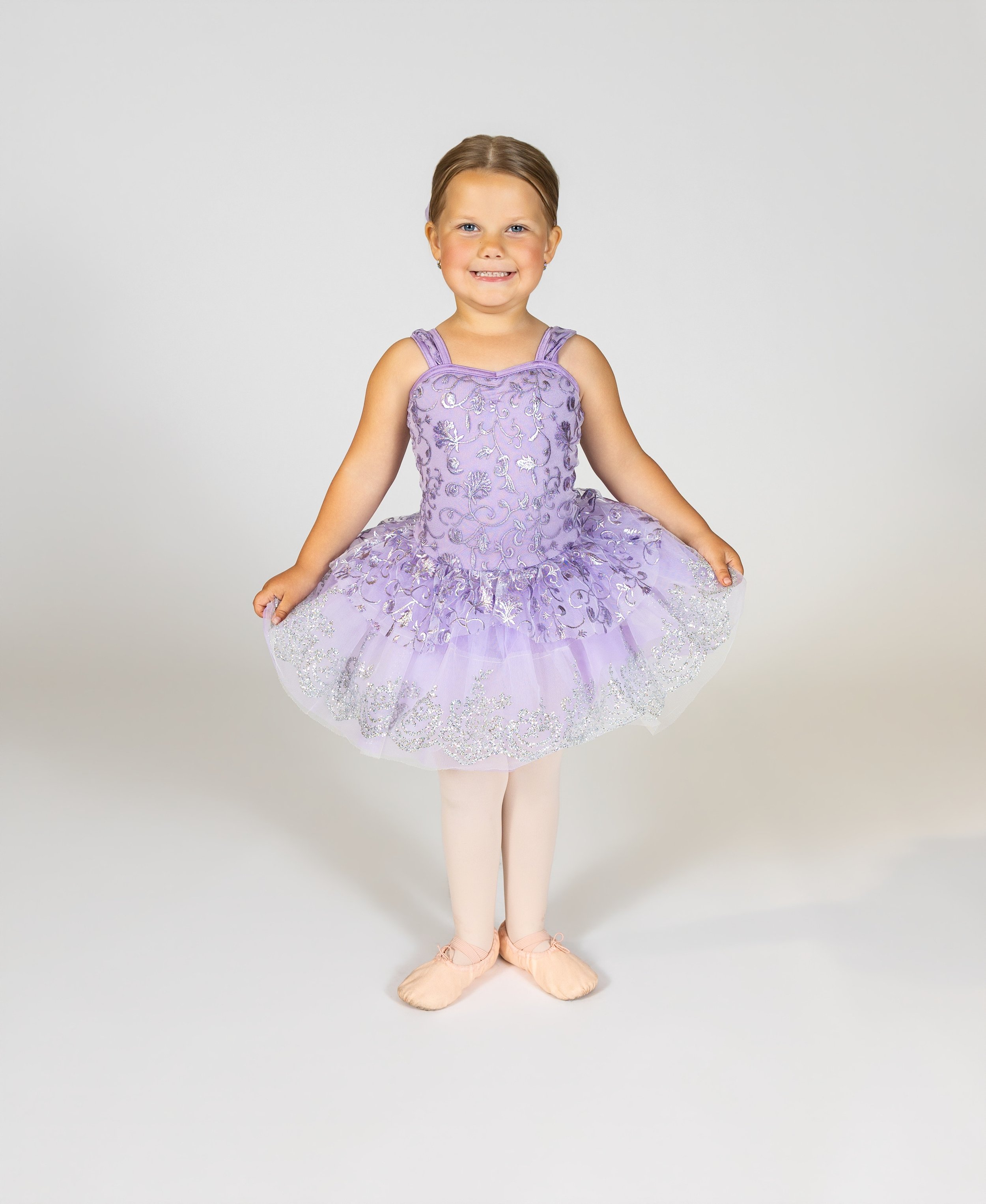 Dancer dressed as a ballerina in a purple tutu with floral embroidery, pink tights, and ballet slippers, standing against a plain gray background and smiling at the camera.