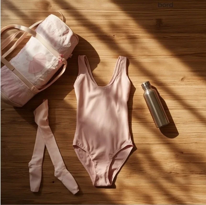 A pink ballet leotard, ballet tights, a tote bag with floral design, and a stainless steel water bottle are arranged on a wooden floor with sunlight streaming across.