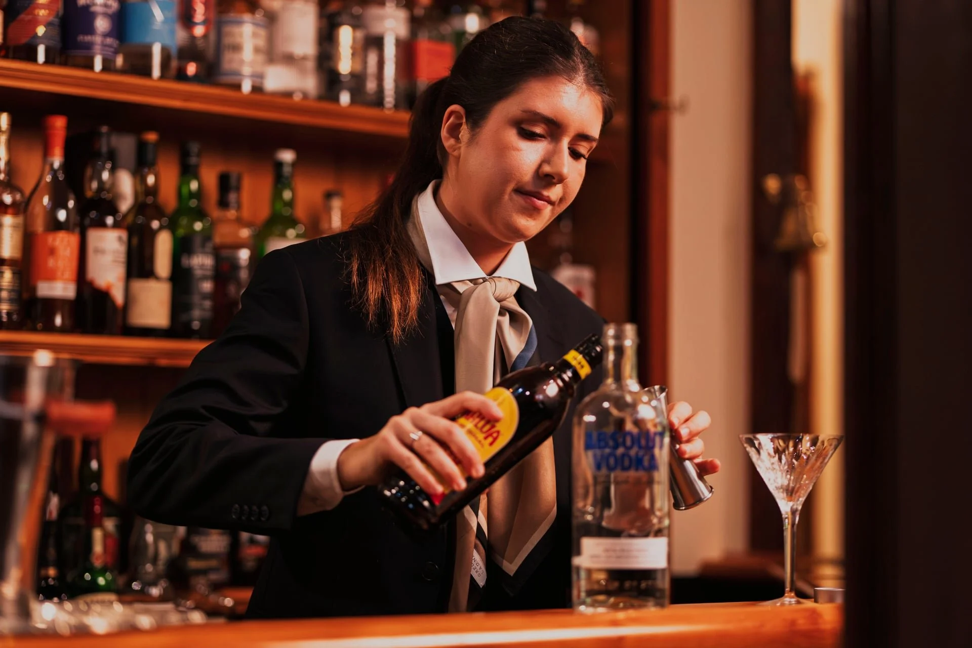 A female bartender in formal attire preparing a cocktail behind a bar with liquor bottles on shelves.