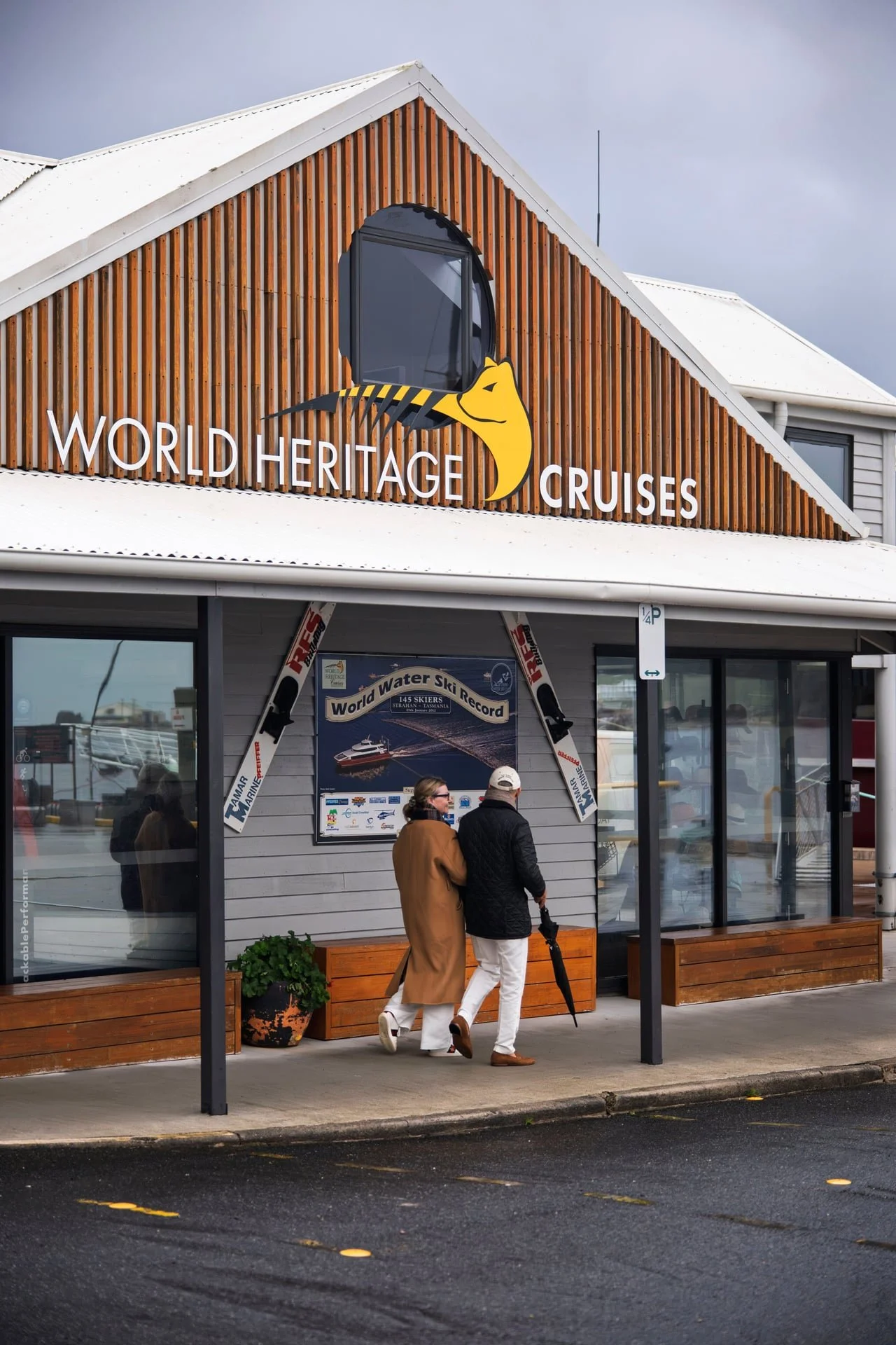 Two women walking past the entrance of a building with a sign reading "World Heritage Cruises." The building has a wooden façade, large glass windows, and a small covered outdoor area. The scene appears to be at a waterfront or marina, with boats visible through the windows and on a sign referencing a world water ski record.