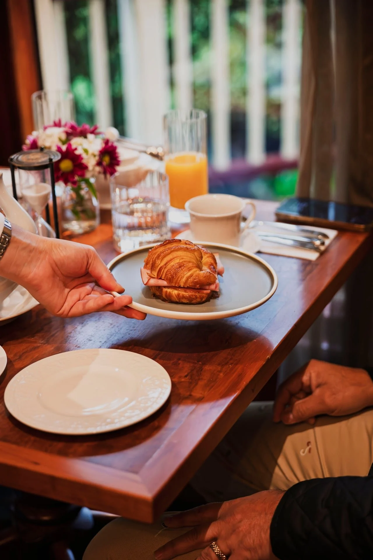 A person is holding a plate with a croissant sandwich at a breakfast table. The table has a glass of orange juice, a cup, and a flower arrangement in the background, with a window showing greenery outside.