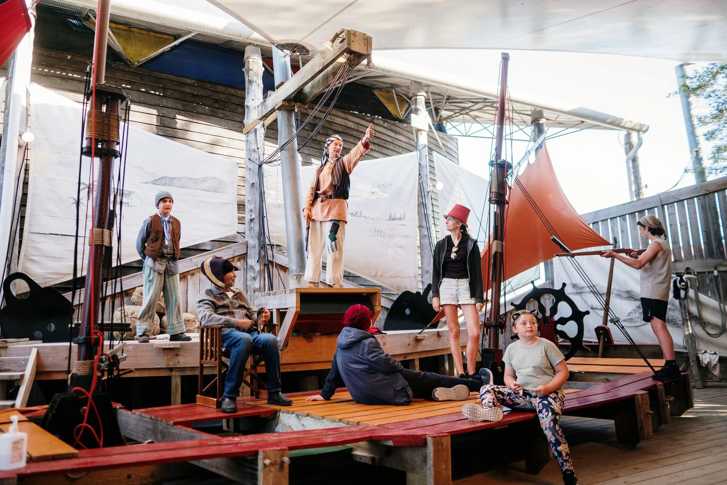 A theatrical performance on a wooden stage with a pirate theme, featuring actors in period costumes with pirate hats, a man in a striped shirt, a woman with a red hat, and children, with nautical props and background scenery resembling a ship's deck.