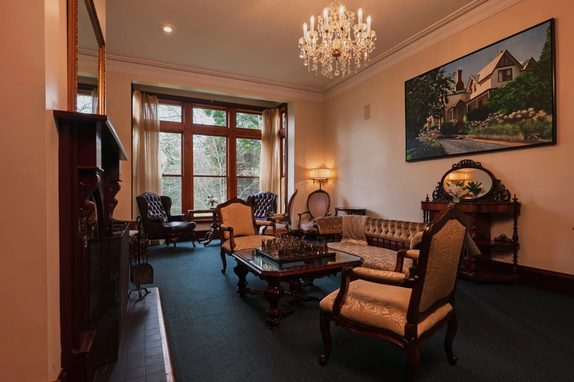 Living room with large window, vintage furniture, chandelier, framed landscape mural, and a blue carpet.
