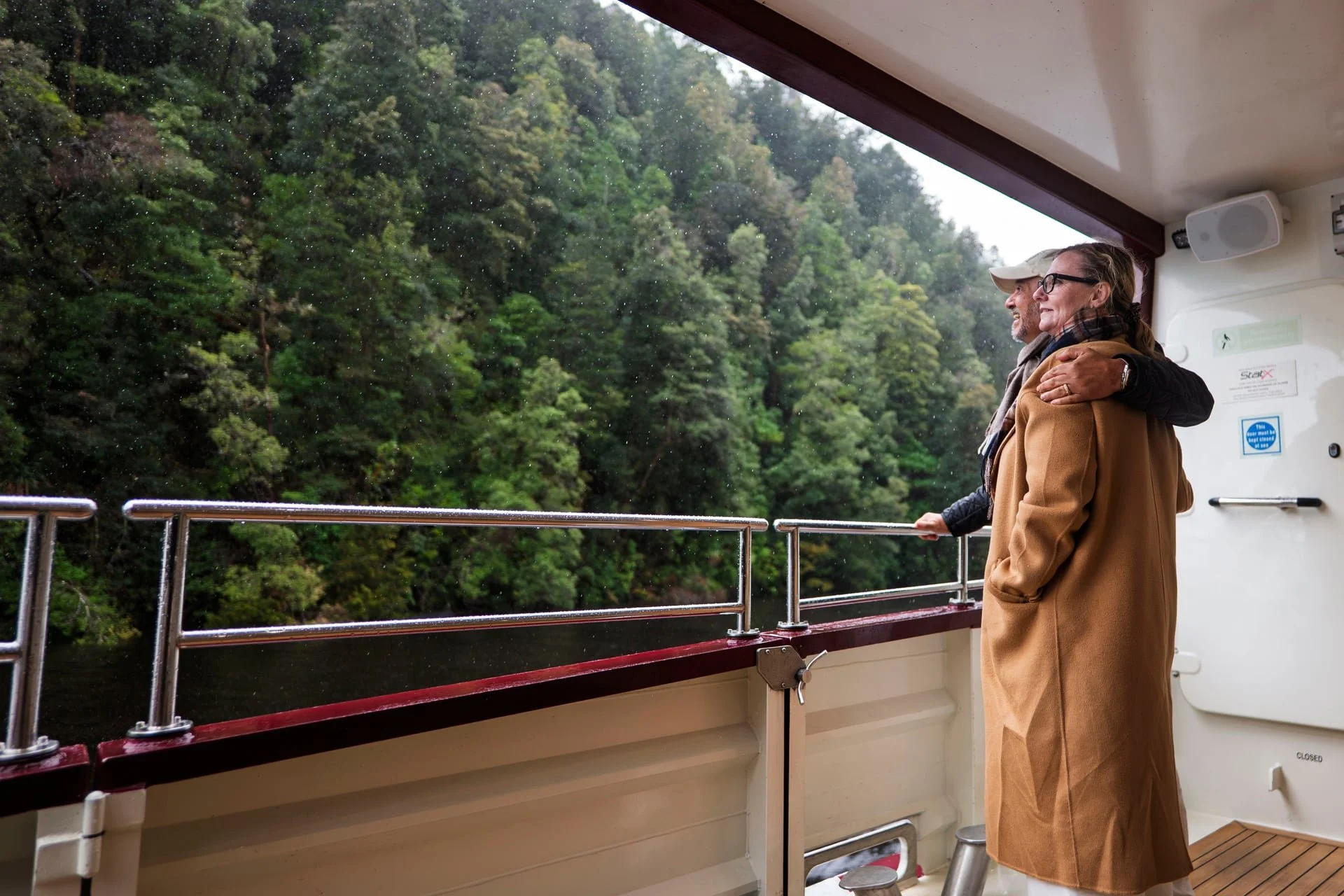 Two people standing on a boat deck looking out at a lush green forested hillside during rain. One person is wearing a camel coat and glasses, and the other is in a cap and dark coat.
