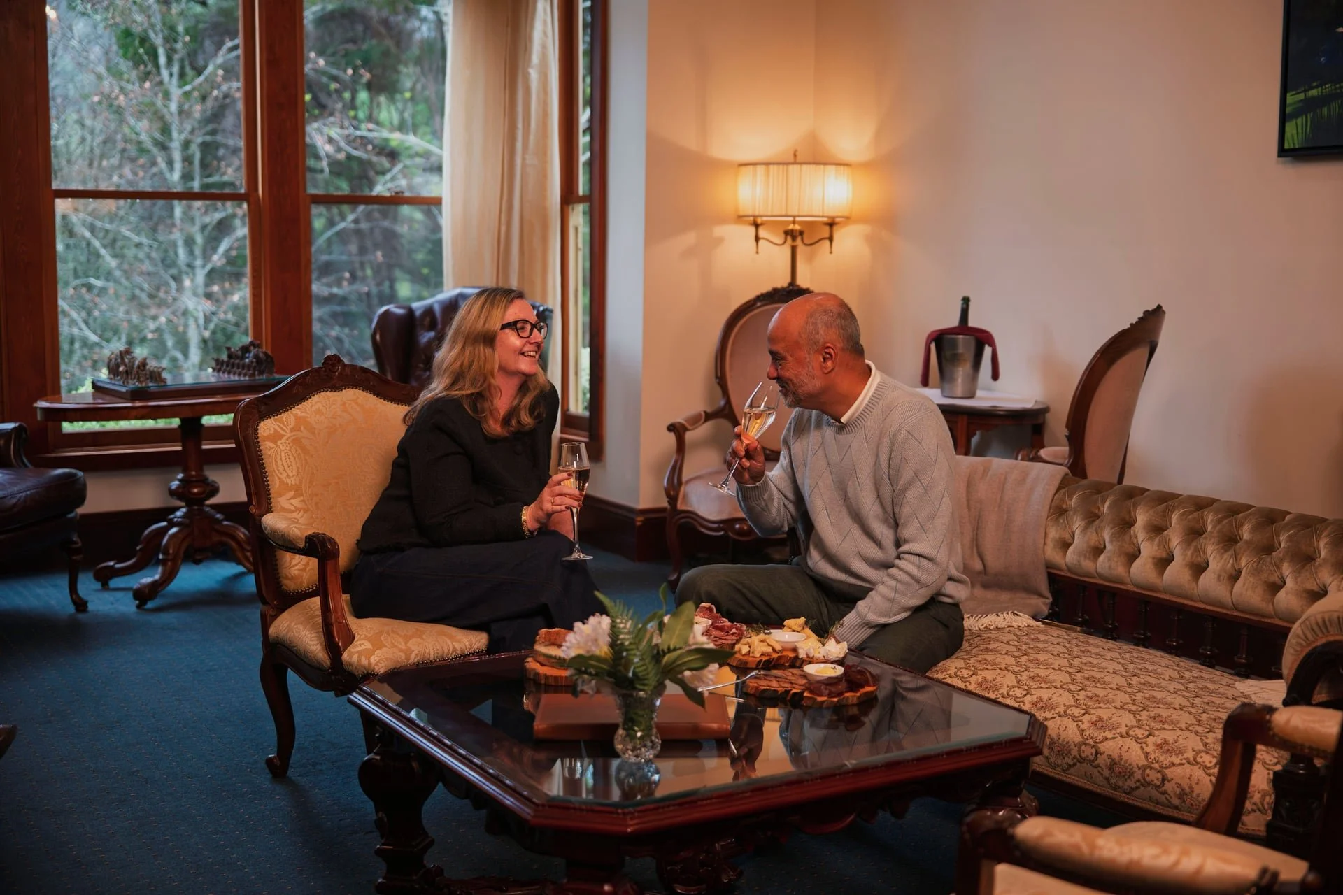 A man and woman sitting on a sofa, smiling and sharing a toast with champagne glasses in a cozy living room with large windows, a table with snacks, and warm lighting.