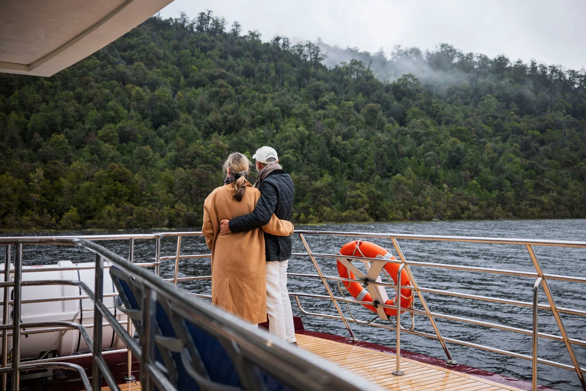 A couple stands on the deck of a boat, embracing while gazing at a lush, green hillside with mist or smoke in the background.