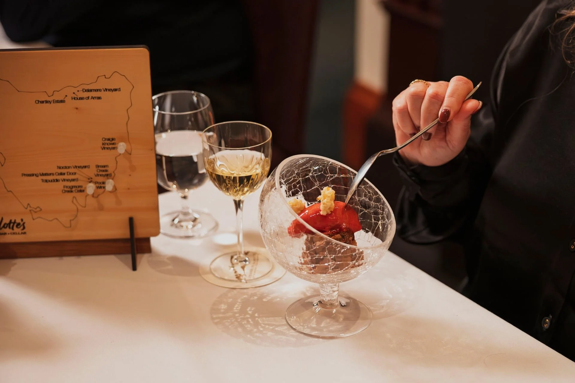 A person using a fork to eat a dessert in a large cracked glass bowl on a white table, with two glasses of white wine and a glass of water nearby, and a wooden menu stand with a map of a vineyard.