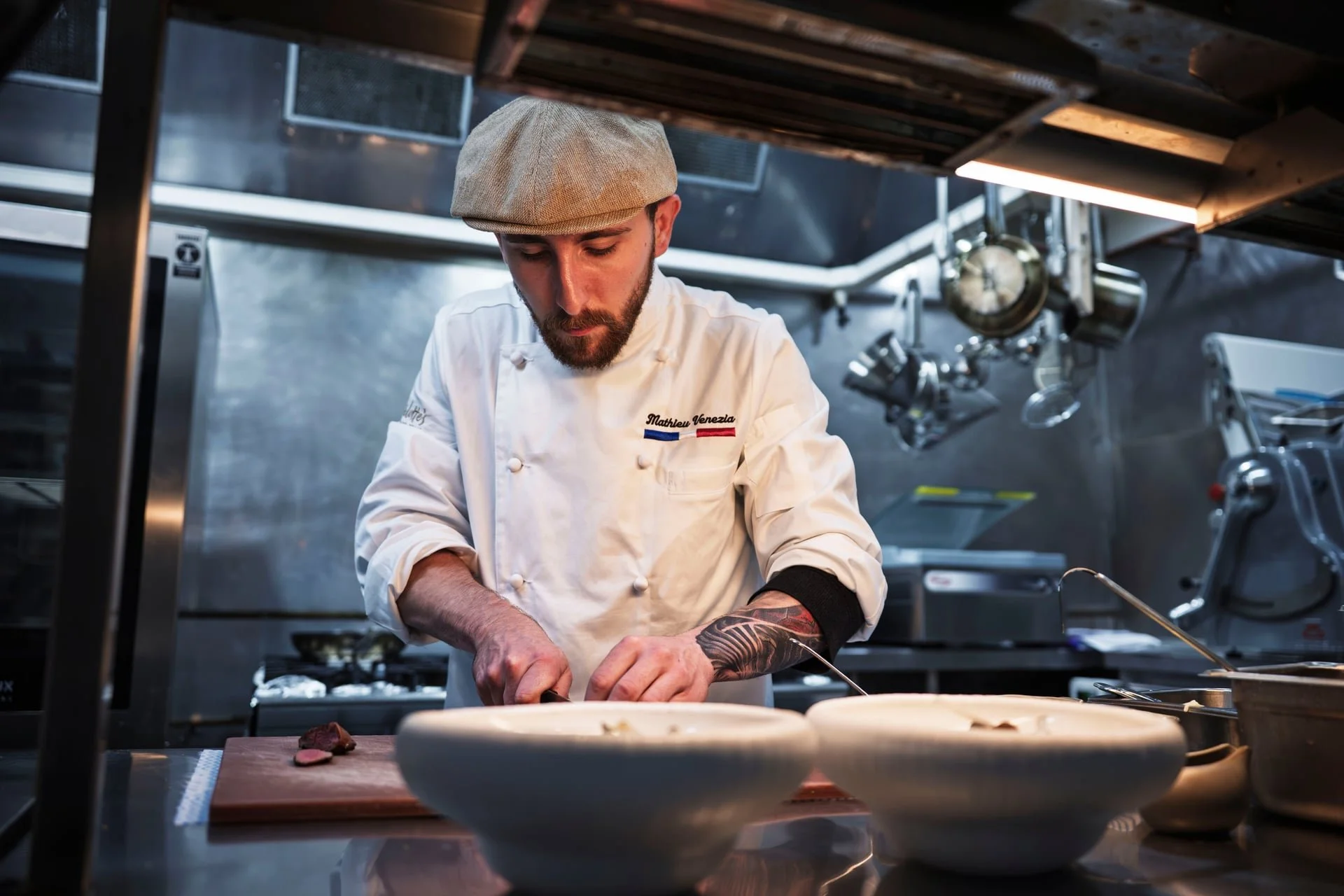 Chef Mathieu preparing a dish in a professional kitchen with hanging pots and pans in the background.