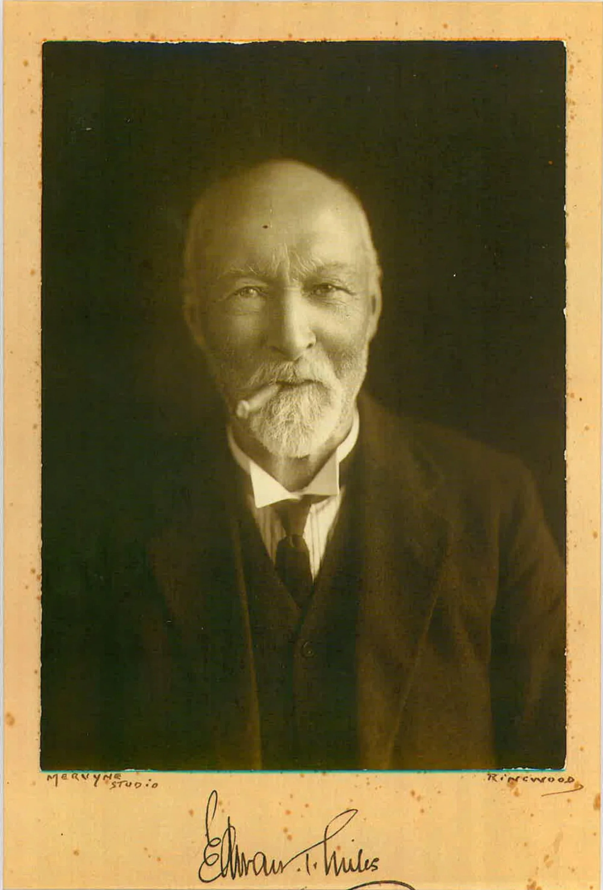 A sepia-toned photograph of an older man with a beard and mustache, dressed in a dark suit, white shirt, and tie, with handwritten text and signatures at the bottom.