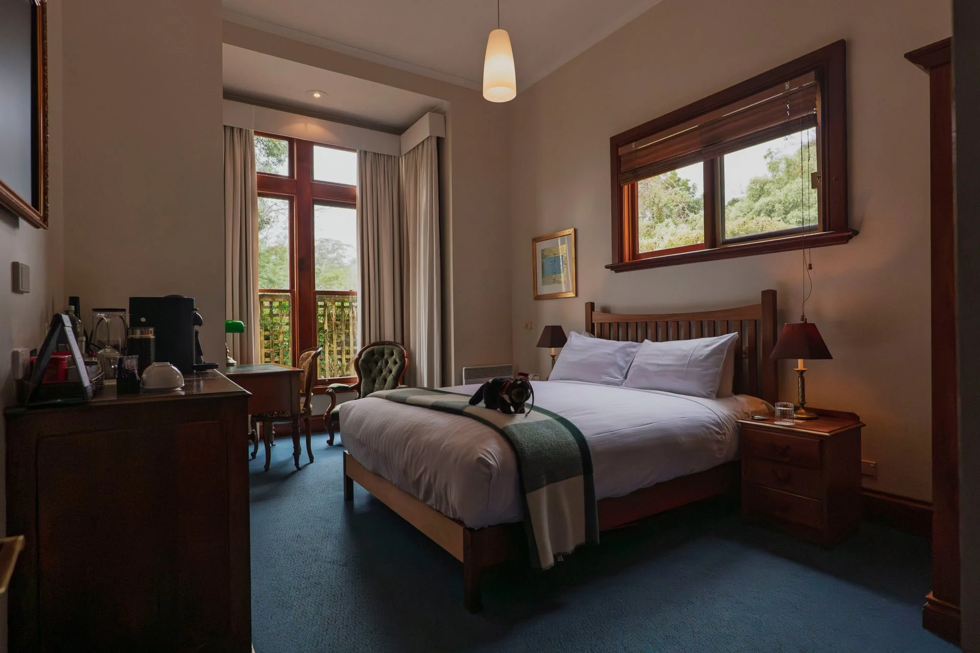 A hotel room with a king-sized bed with white linens, two bedside tables with lamps, a framed picture on the wall, a window above the bed, and a balcony door with curtains to the left. There is a green armchair and a small table near the balcony door, and a wooden desk with a coffee maker and mugs on the left side of the room.