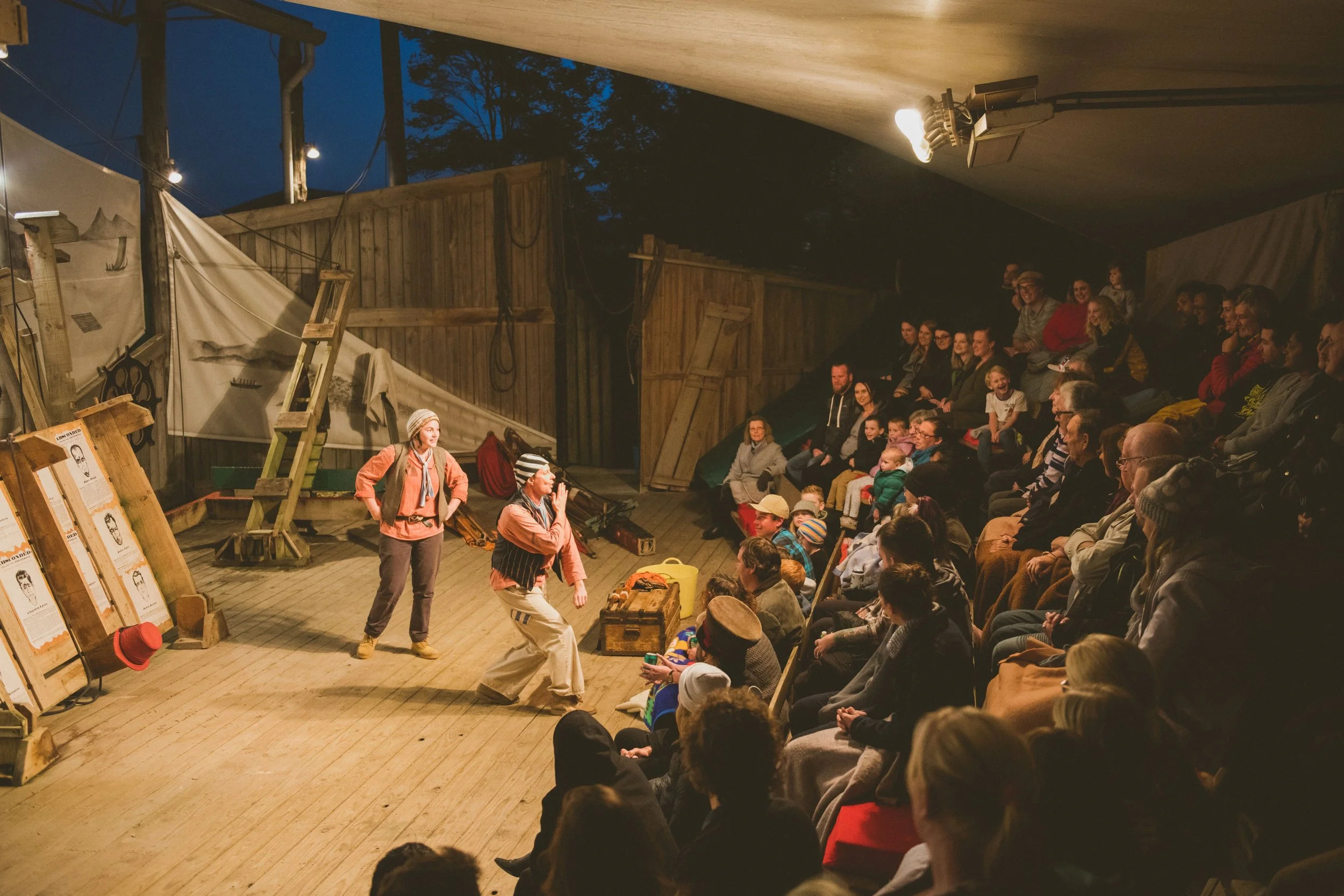 People performing a puppet show on a wooden stage with an audience seated in a semi-circle, set in a rustic outdoor or barn-like environment at night.