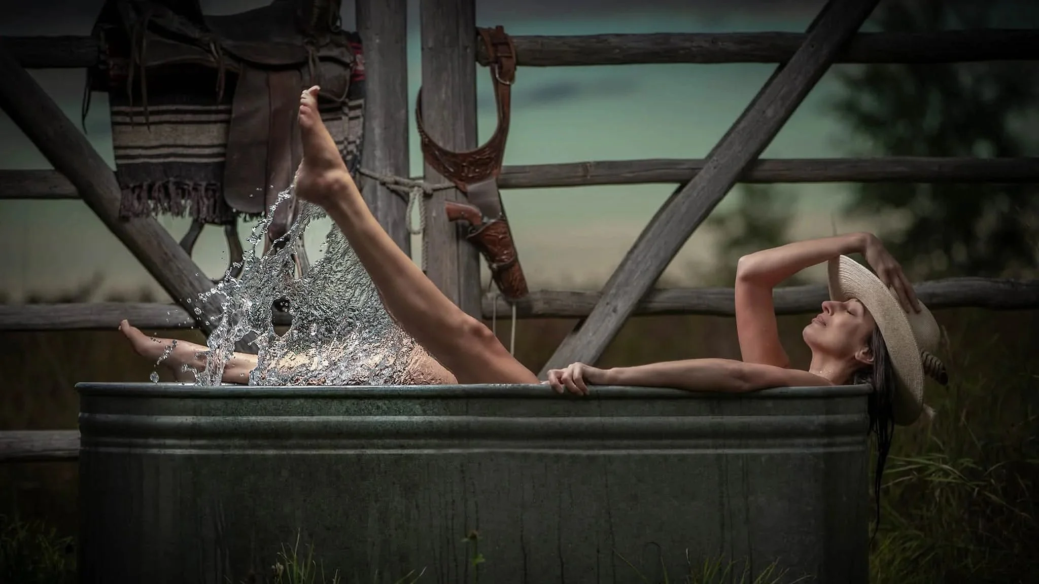 A woman relaxing in a metal bathtub outdoors, wearing a straw hat, with water splashing as she kicks her legs. She is lying back with a serene expression, amidst rustic wooden fencing and green foliage in the background.