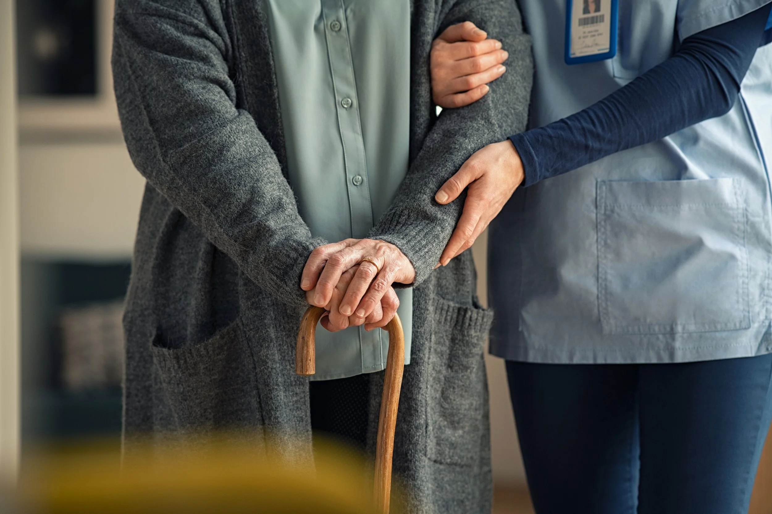 A healthcare worker assists an elderly person with a cane, offering support by holding their arm, in a medical setting.