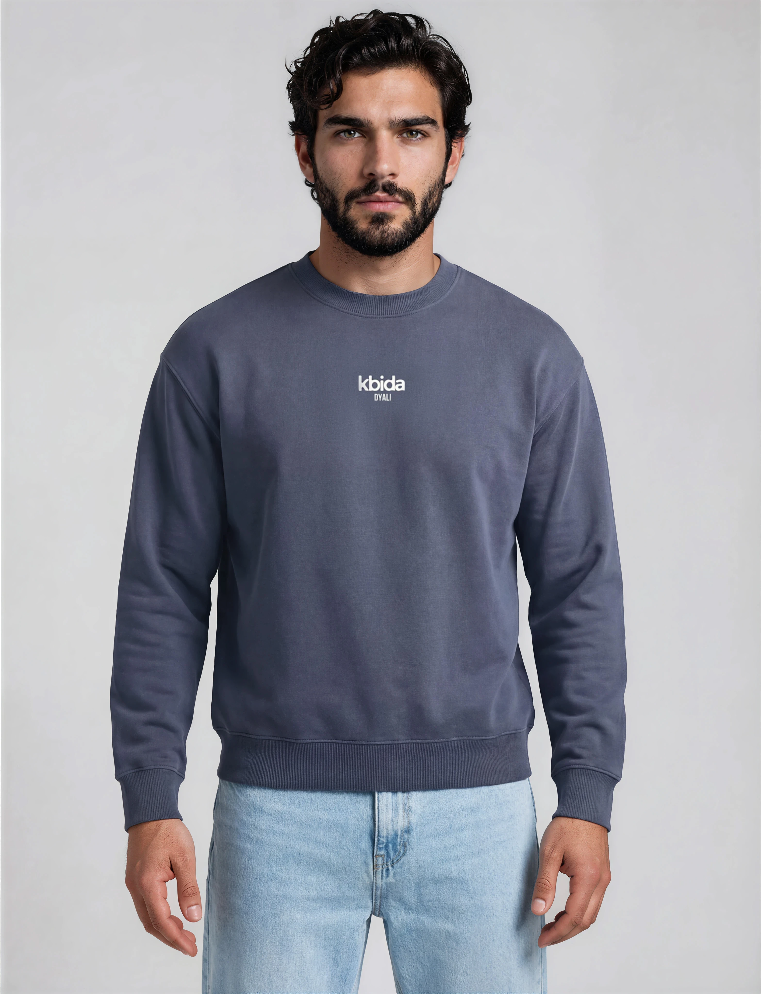 A young man with dark curly hair, beard, wearing a gray sweatshirt with the logo 'kbida DYALI' on the chest, standing against a plain light background.