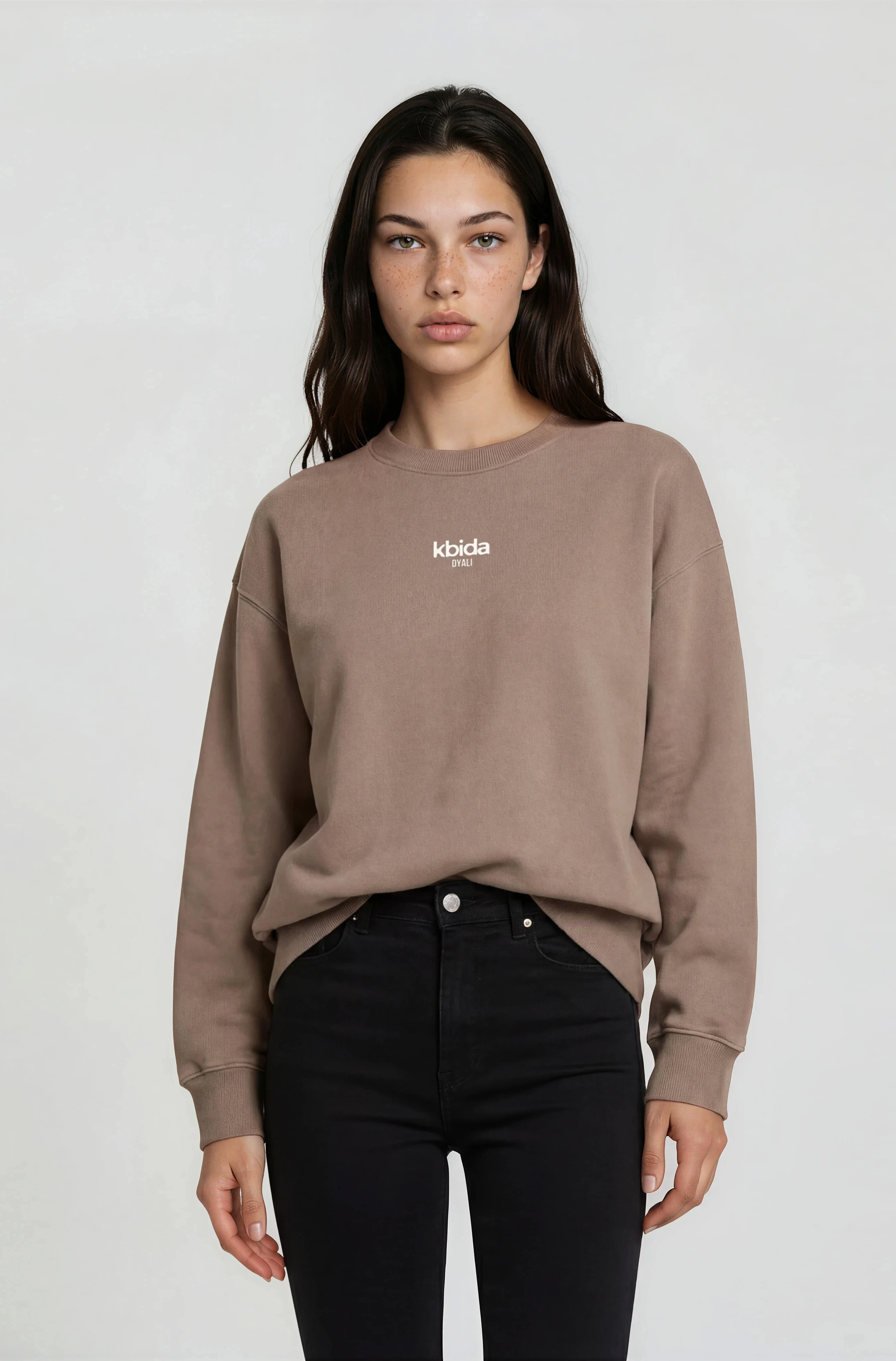 A young woman with dark brown, wavy hair and freckles, wearing a beige sweatshirt with the words 'kbida DYALI' on the front, and black pants, standing against a plain white background.