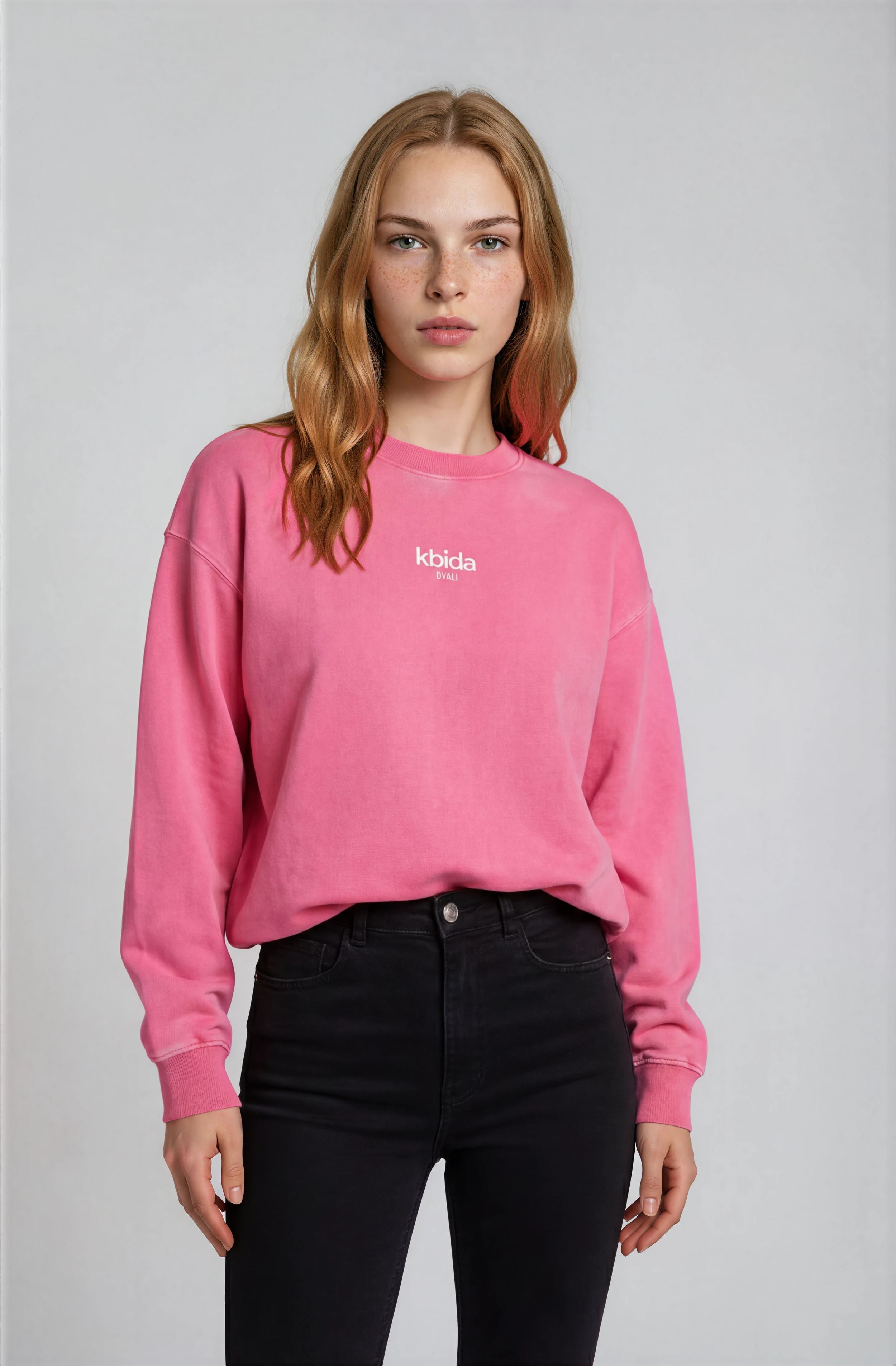 A woman with long wavy red hair, wearing a pink sweatshirt with the words 'kbida DVALI' on the front, and black jeans, standing against a plain light gray background.