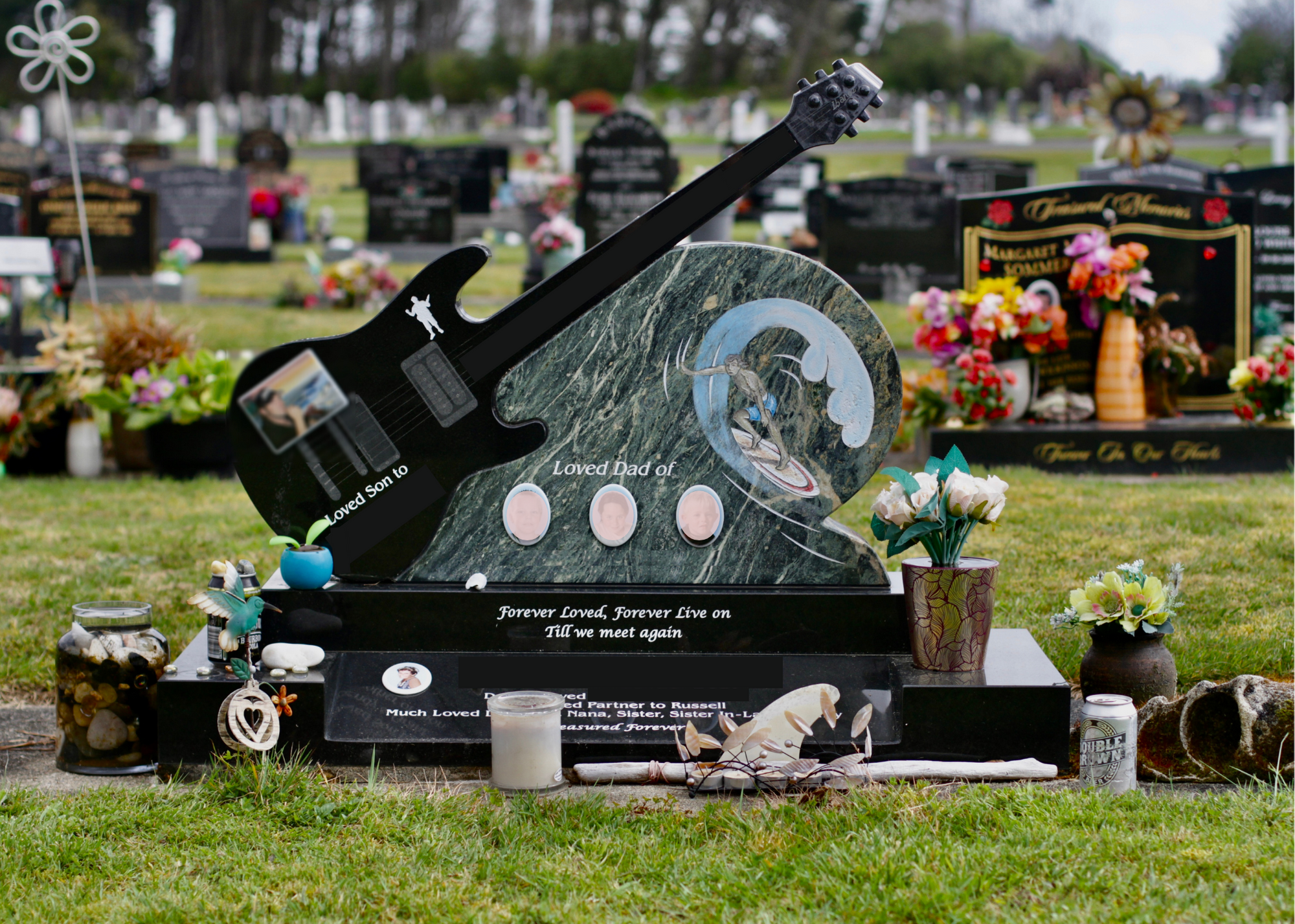 A memorial stone at a cemetery dedicated to a loved father and son, featuring a guitar-shaped plaque with a mountain and surfer painting, flowers, candles, and decorative items.