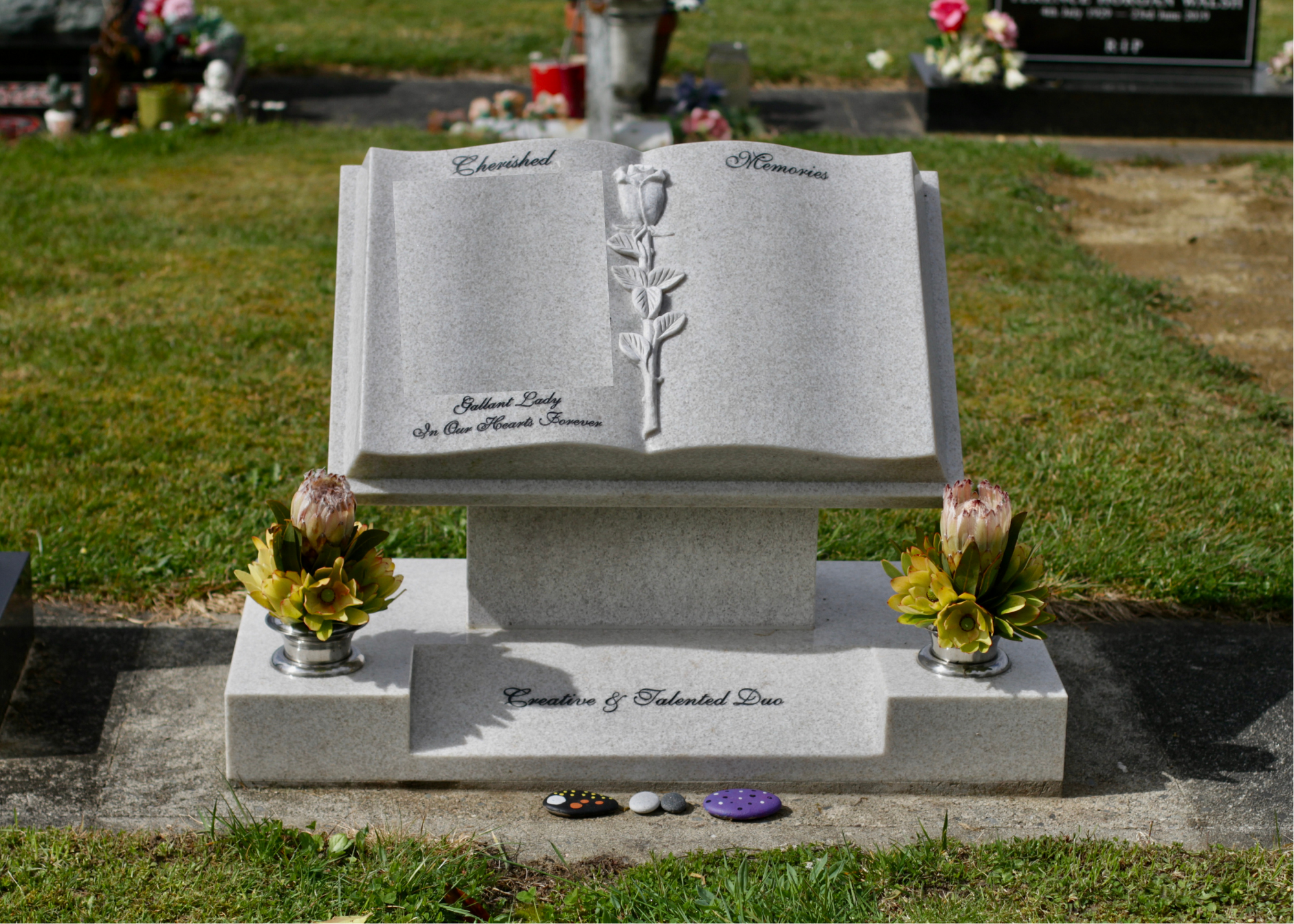 A granite headstone in a cemetery with the words 'Cherished Memories' and 'Gallant Lady In Our Hearts Forever' engraved on it. The headstone is decorated with a carved rose and two small flower arrangements at the base. Three painted rocks are placed in front of it, one black with multicolored circles, one white, and one purple with white polka dots.