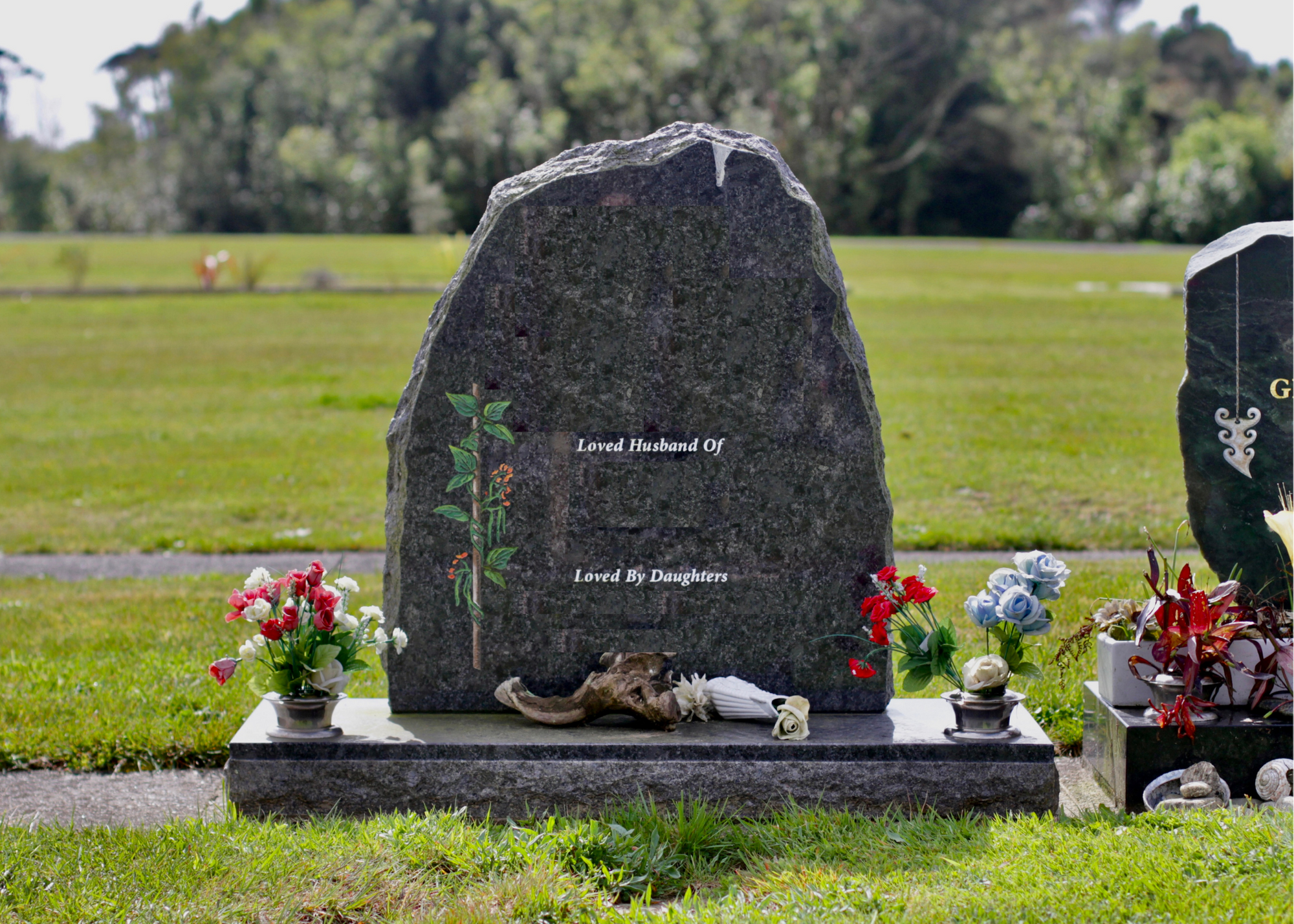 A black granite headstone in a cemetery with engraved writing, surrounded by colorful artificial flowers and a driftwood piece, with green grass and trees in the background.