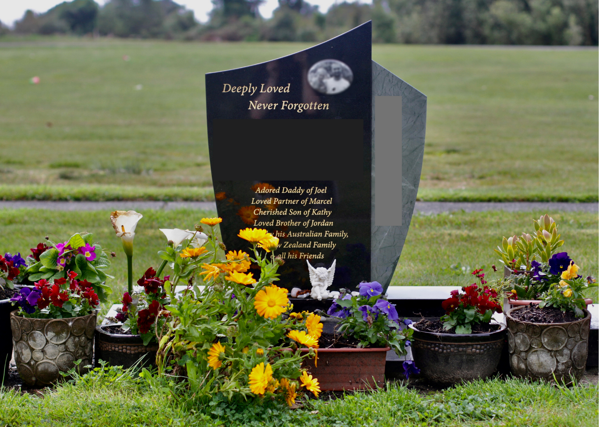 Gravestone with colorful flowers in pots around it at a cemetery, with green grass and trees in the background.