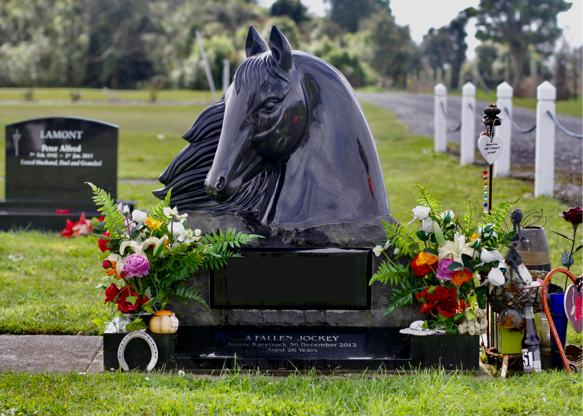 Gravestone with black horse head sculpture, flowers, and memorial items in a cemetery.