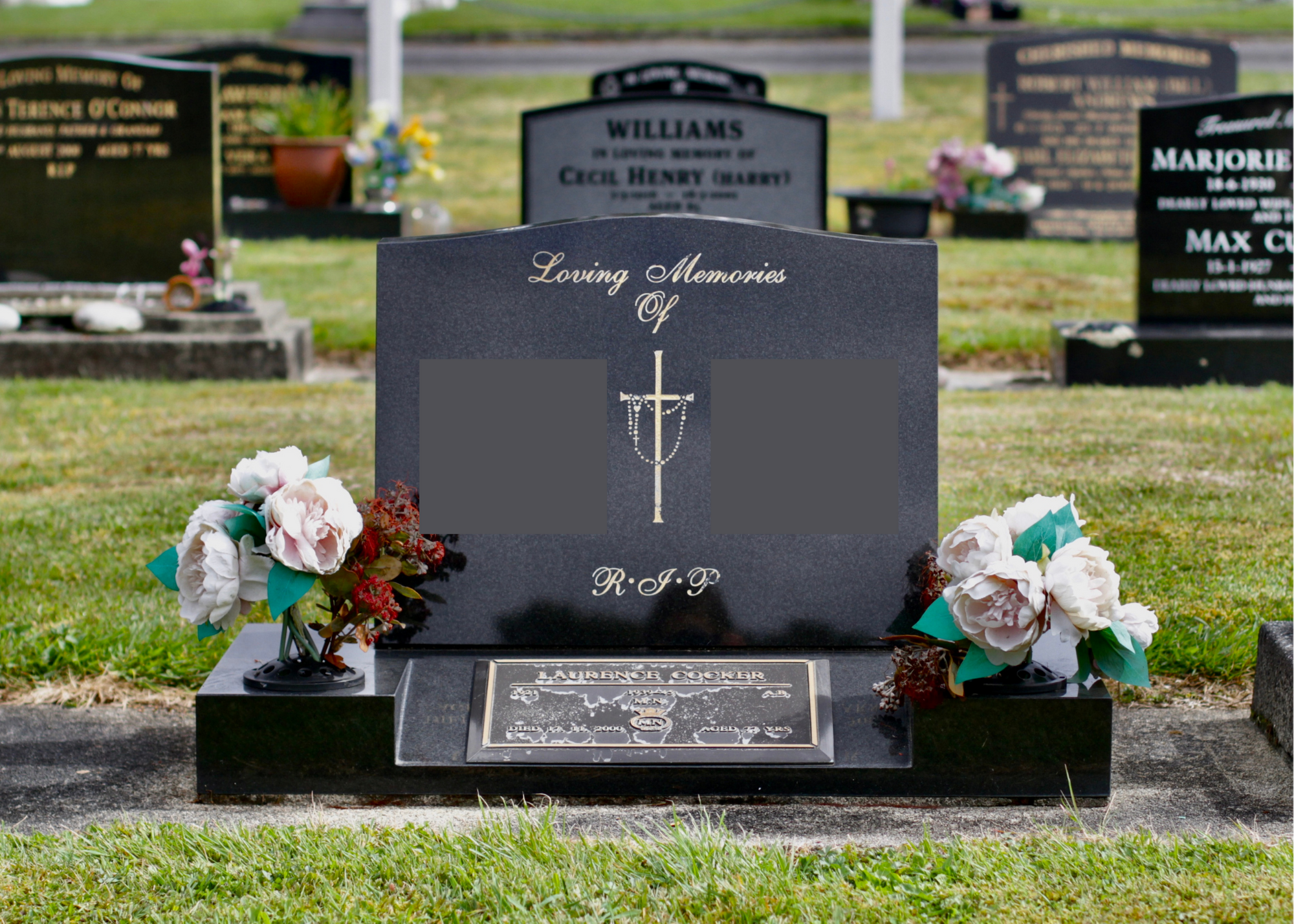 A black granite headstone at a cemetery with engraved memorial text and a small plaque. The headstone has floral arrangements on either side and is surrounded by other similar graves in the background.