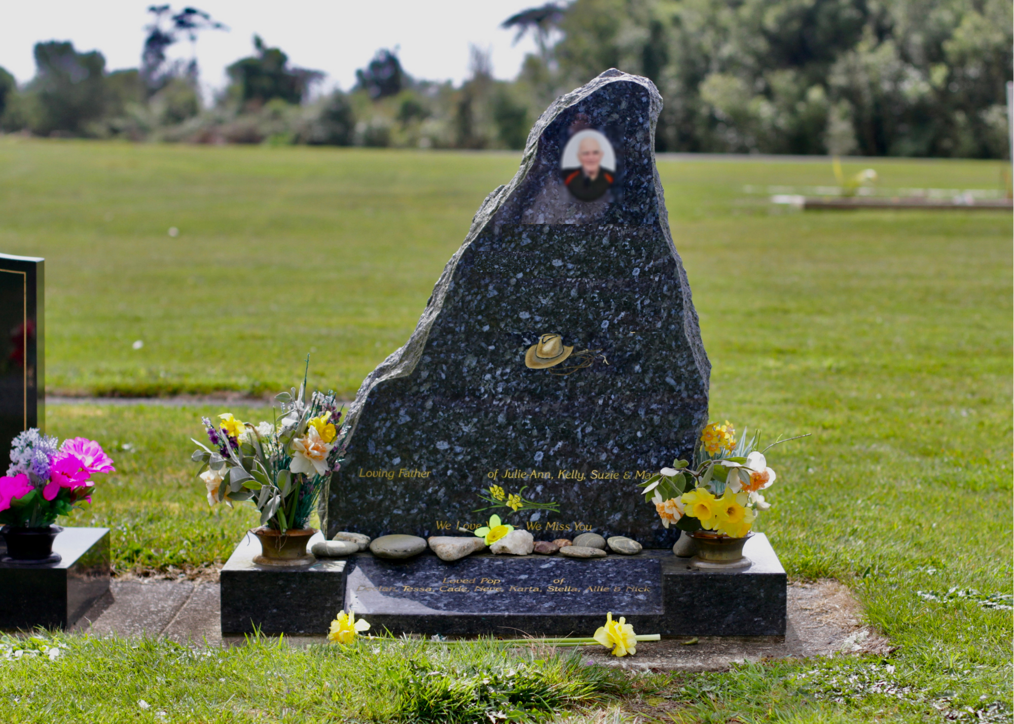 A polished black granite headstone in a cemetery, decorated with artificial flowers, small rocks, and a photo of an elderly woman at the top. The headstone has inscriptions honoring a caring father and loved ones who have passed away.