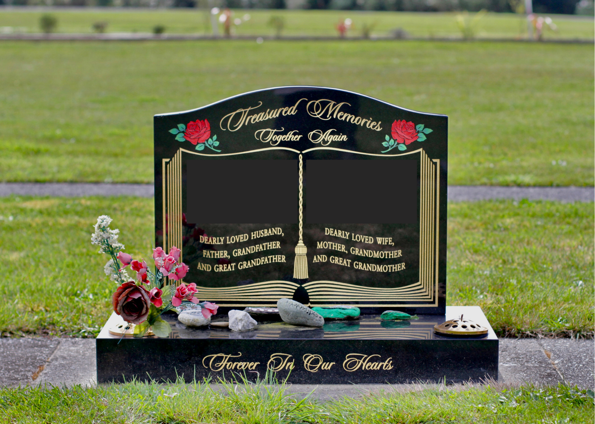 Black granite headstone with engraved gold text and floral decorations, surrounded by flowers and stones, located in a grassy cemetery.