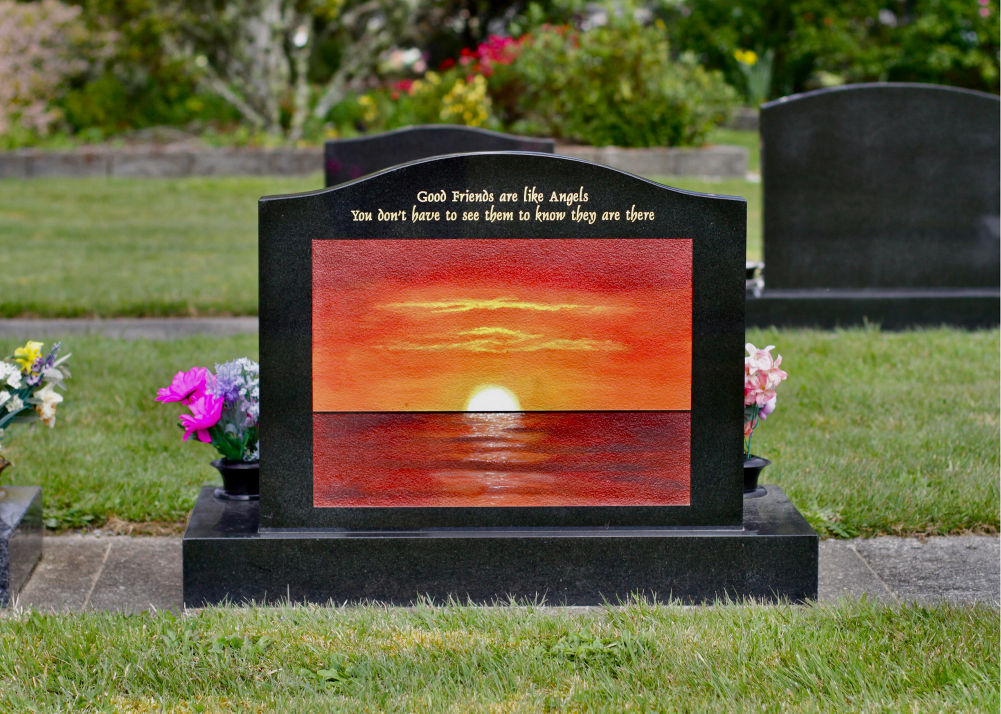 Gravestone with a sunset painting and the quote 'Good friends are like angels. You don't have to see them to know they are there' inscribed on it, surrounded by flowers in a cemetery.