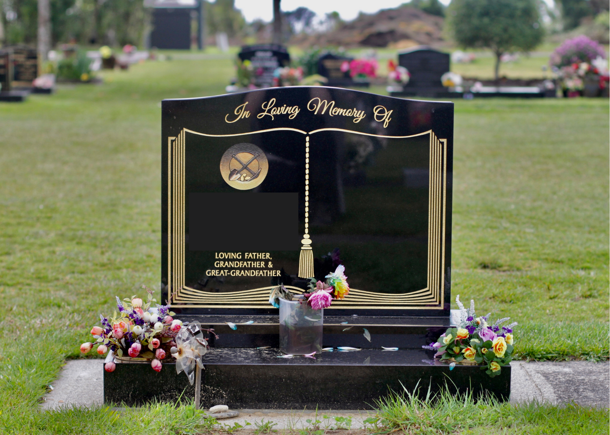 A black granite funeral headstone with gold embossed text and decorations, surrounded by flowers at a cemetery.