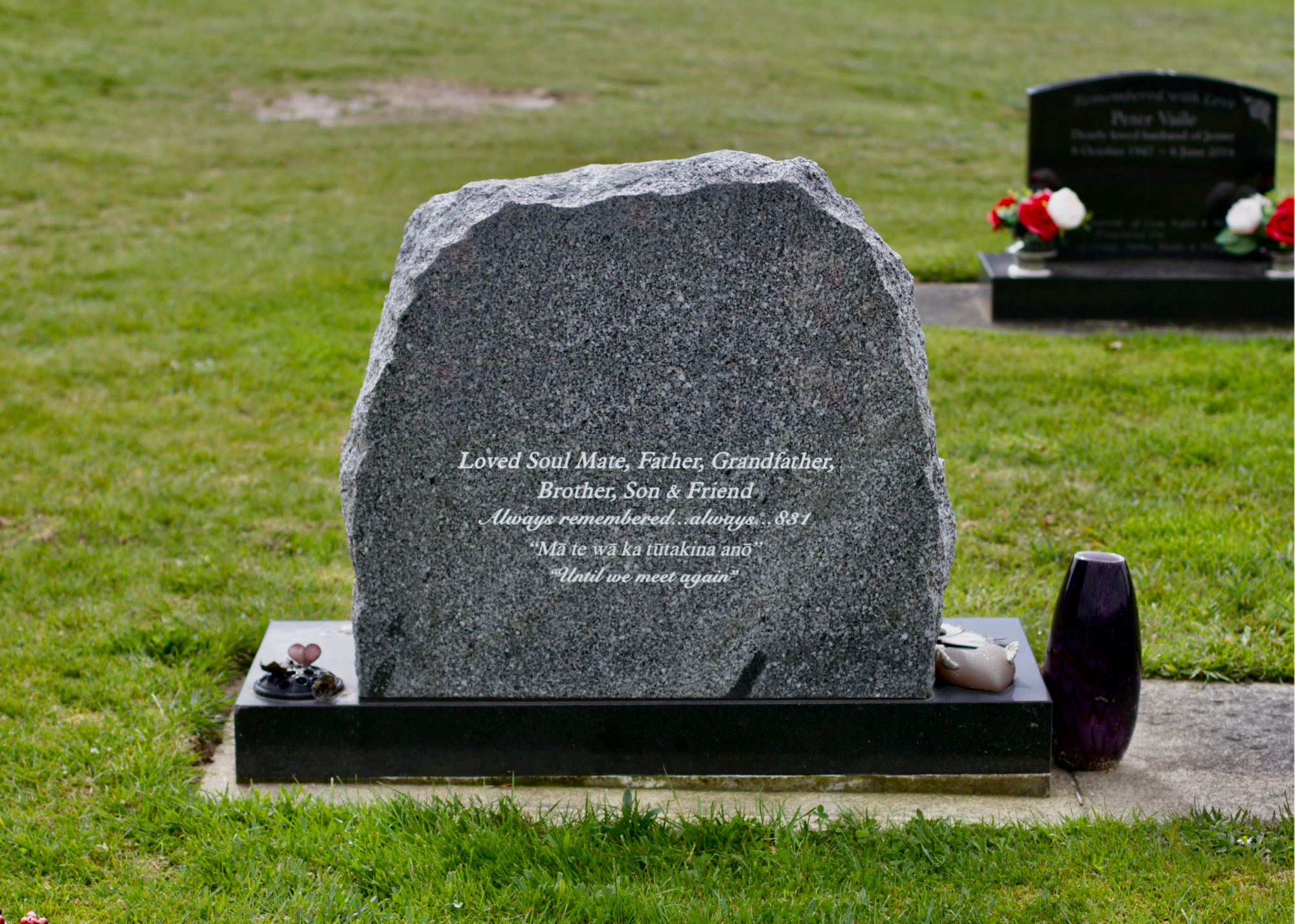 Gray granite headstone in a cemetery with an inscription in English and Hawaiian, surrounded by green grass and a purple vase to the right.