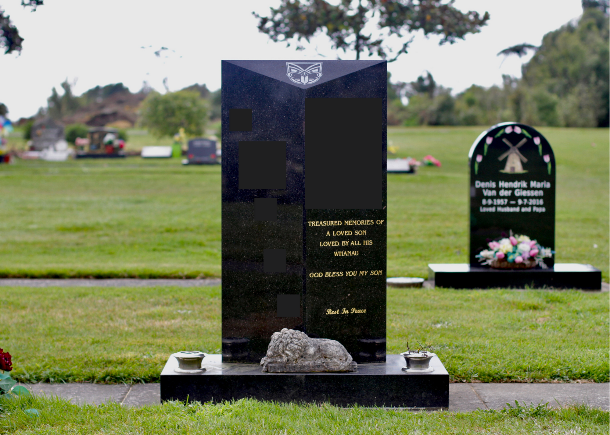 A black granite tombstone with a carved lion at the base, in a cemetery with other graves and a grassy landscape in the background.