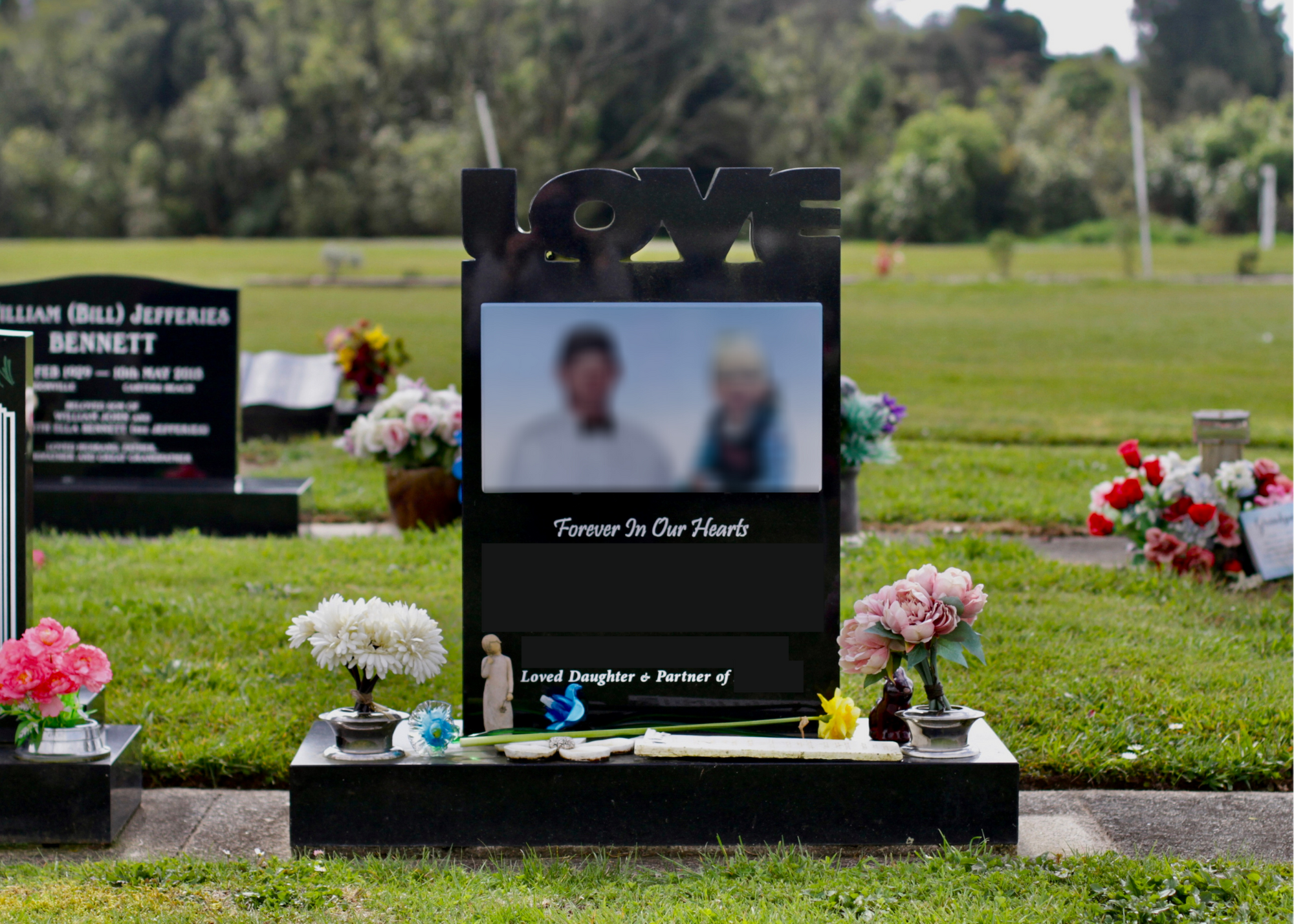 Grave with a black headstone decorated with flowers and a photograph at a cemetery. The headstone has the word 'LOVE' on top and the phrase 'Forever In Our Hearts.' There are various colorful flowers and small statues placed around the grave.