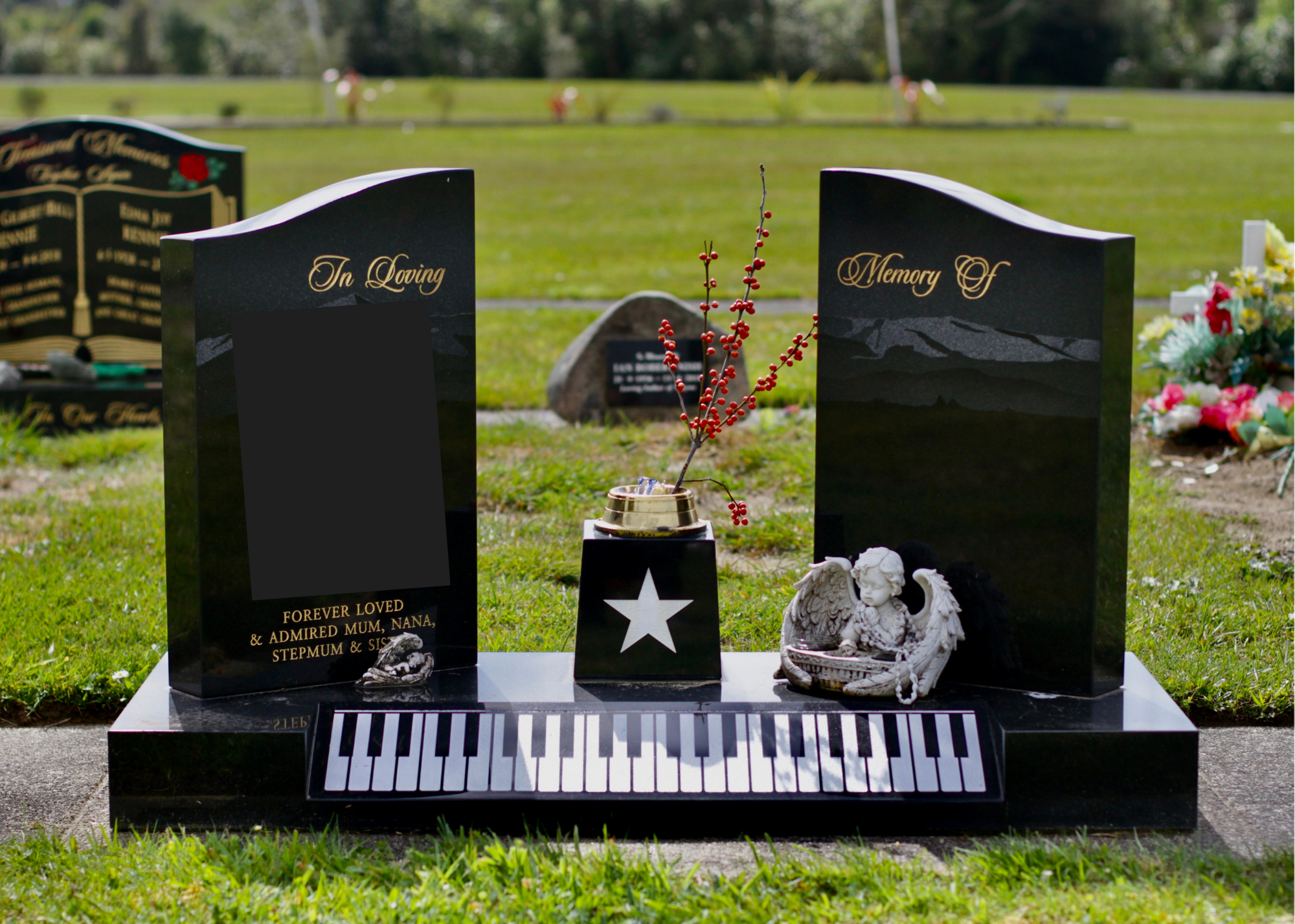 Black granite gravestone with plaques engraved 'In Loving Memory of' and 'Memory of', flowers in a small pot, a angel figurine, and a digital keyboard design at the base, in a cemetery with other headstones and green grass.