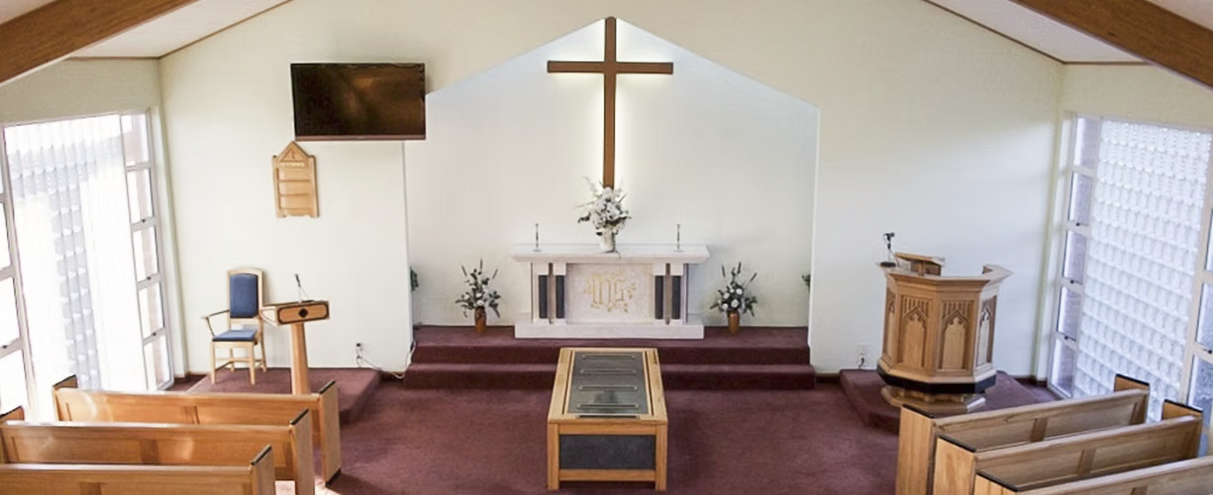 Interior of a Hagedorns Buller Funeral Services chapel, a small church with a central altar, a wooden cross above it, and an empty pulpit on the right. The church has large windows on both sides and wooden pews.