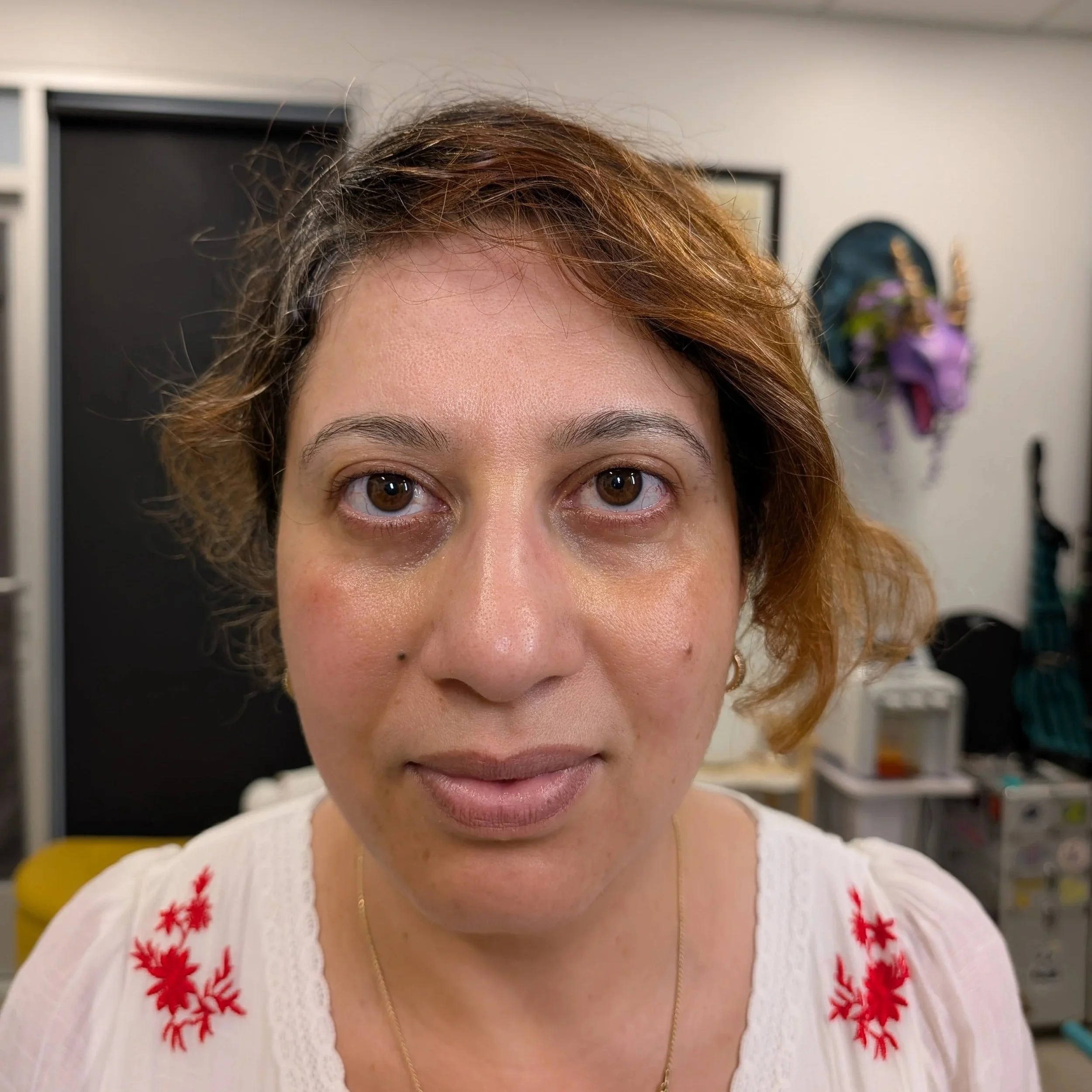 Close-up of a woman's face, showing her forehead, eyes, and part of her nose, with curly reddish-brown hair and wearing a white blouse with red embroidery on the shoulders, indoors with a blurred background. before microbladed brows