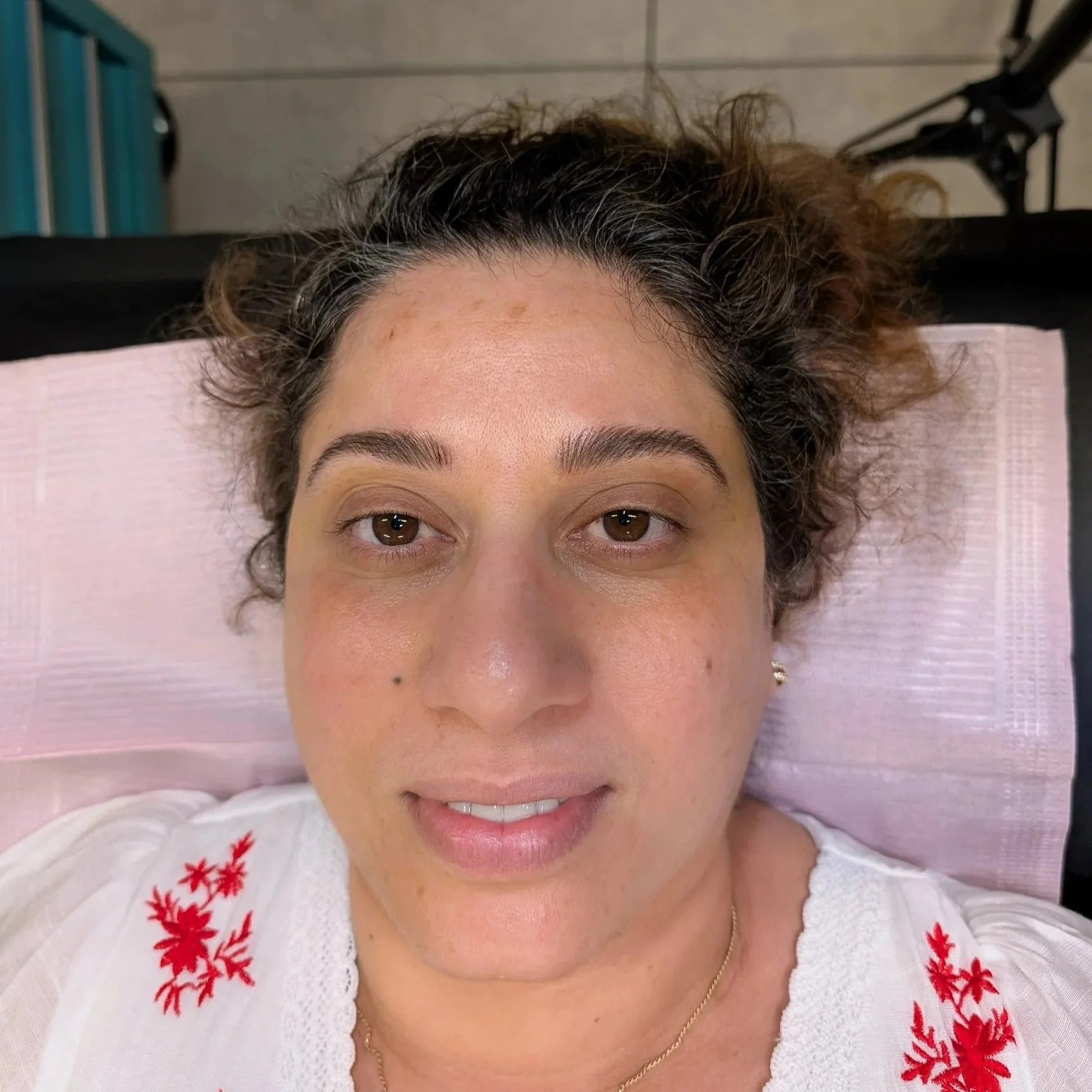 Close-up of a woman lying on a pink pillow, smiling with her eyes open, wearing a white top with red embroidery, and a gold chain necklace. after microbladed brows