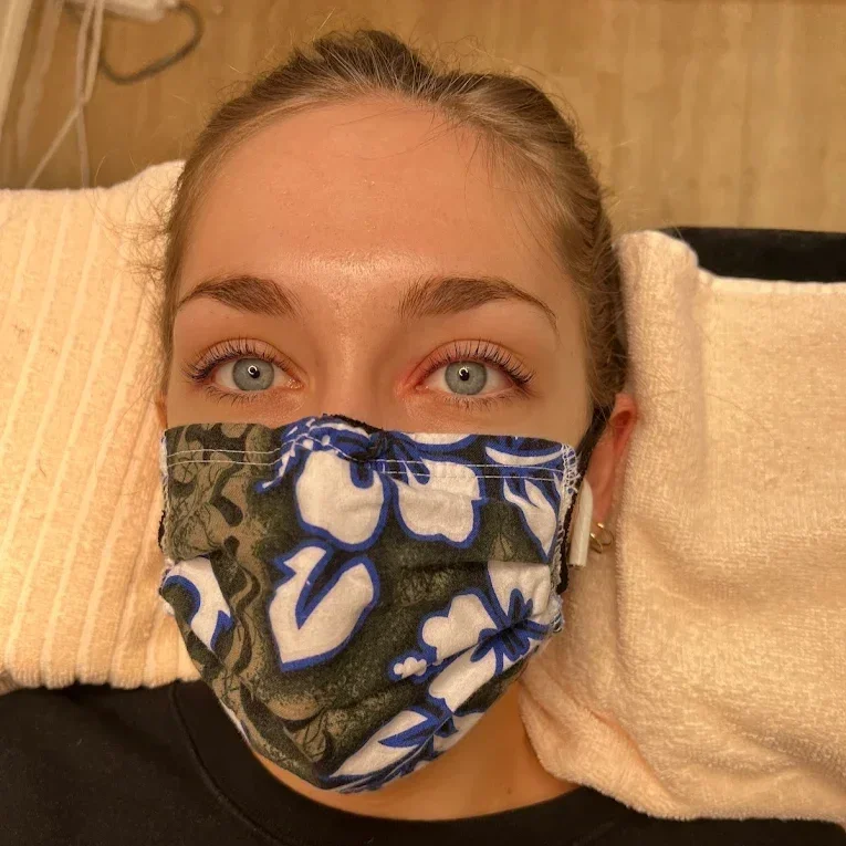 Close-up of a woman with blue eyes and eyebrows wearing a patterned face mask, lying on a beige pillow in a room with brown walls. korean lash lift
