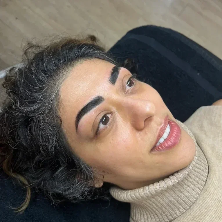 Close-up of a woman with curly hair and makeup, lying down on a black surface, wearing a beige turtleneck sweater, looking slightly to the side. brow lamination