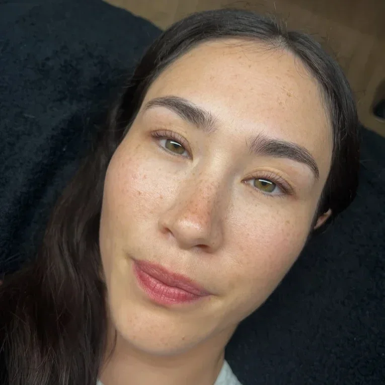 Close-up of a young woman with freckles, brown hair, and hazel eyes, lying on a dark surface and looking at the camera. korean lash lift