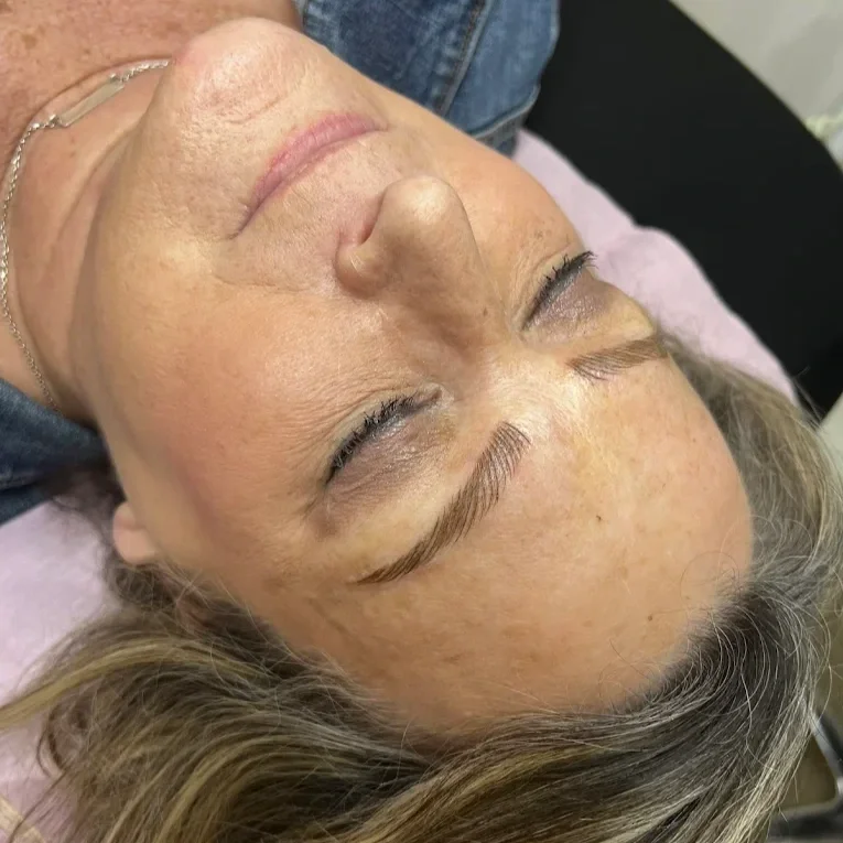 Close-up of an older woman lying down with her eyes closed, showing her face and part of her upper body, including her hair and a pink cloth underneath her head. microbladed brows