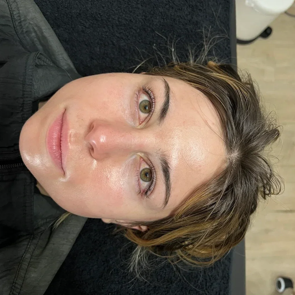 Close-up of a woman with fair skin and brown hair with blonde highlights, lying down on a black surface, looking at the camera with green eyes. She has minimal makeup and a natural expression. lash lift
