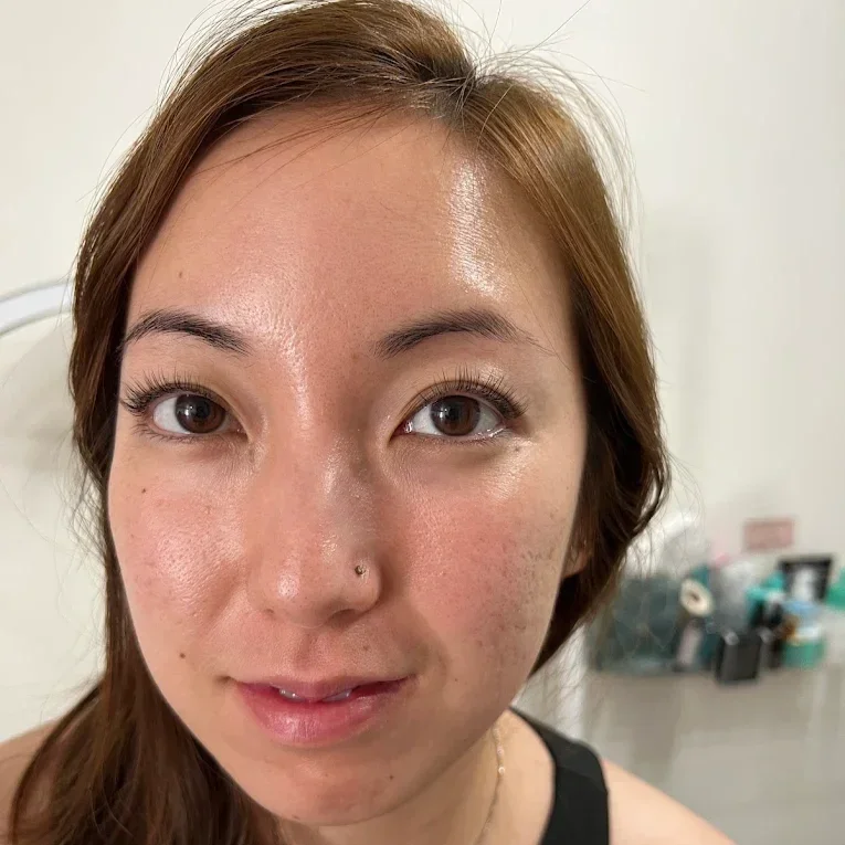 Close-up of a young woman with light skin, brown hair, brown eyes, and a nose piercing, looking at the camera, in a room with a white wall and some objects blurred in the background. korean lash lift