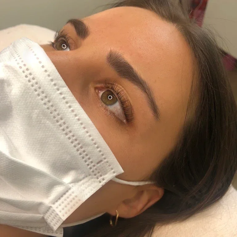 Close-up of a woman wearing a white face mask, lying down, with her eyes open and looking upwards. lash lift