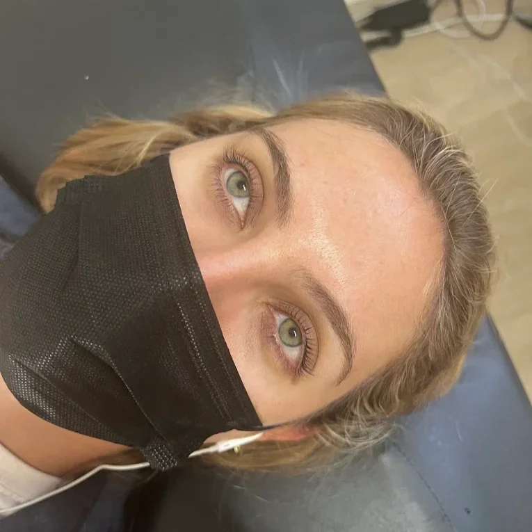 Close-up of a woman lying on a black surface, wearing a black face mask, with blonde hair, light-colored eyes, and well-groomed eyebrows. lash lift