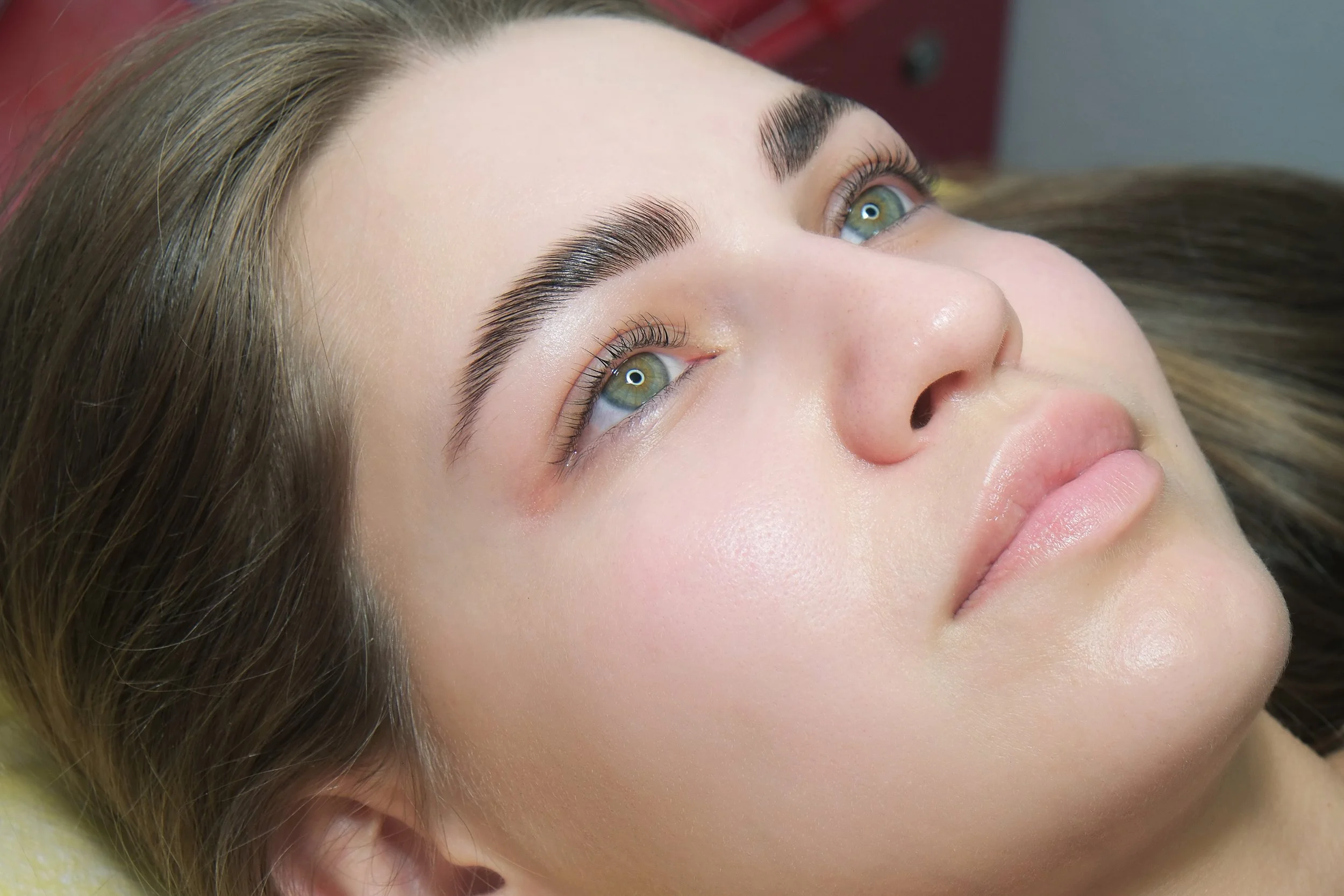 Close-up of a young woman with long brown hair lying down, her face turned slightly upward, showcasing their well-groomed eyebrows, natural makeup, and smooth skin.