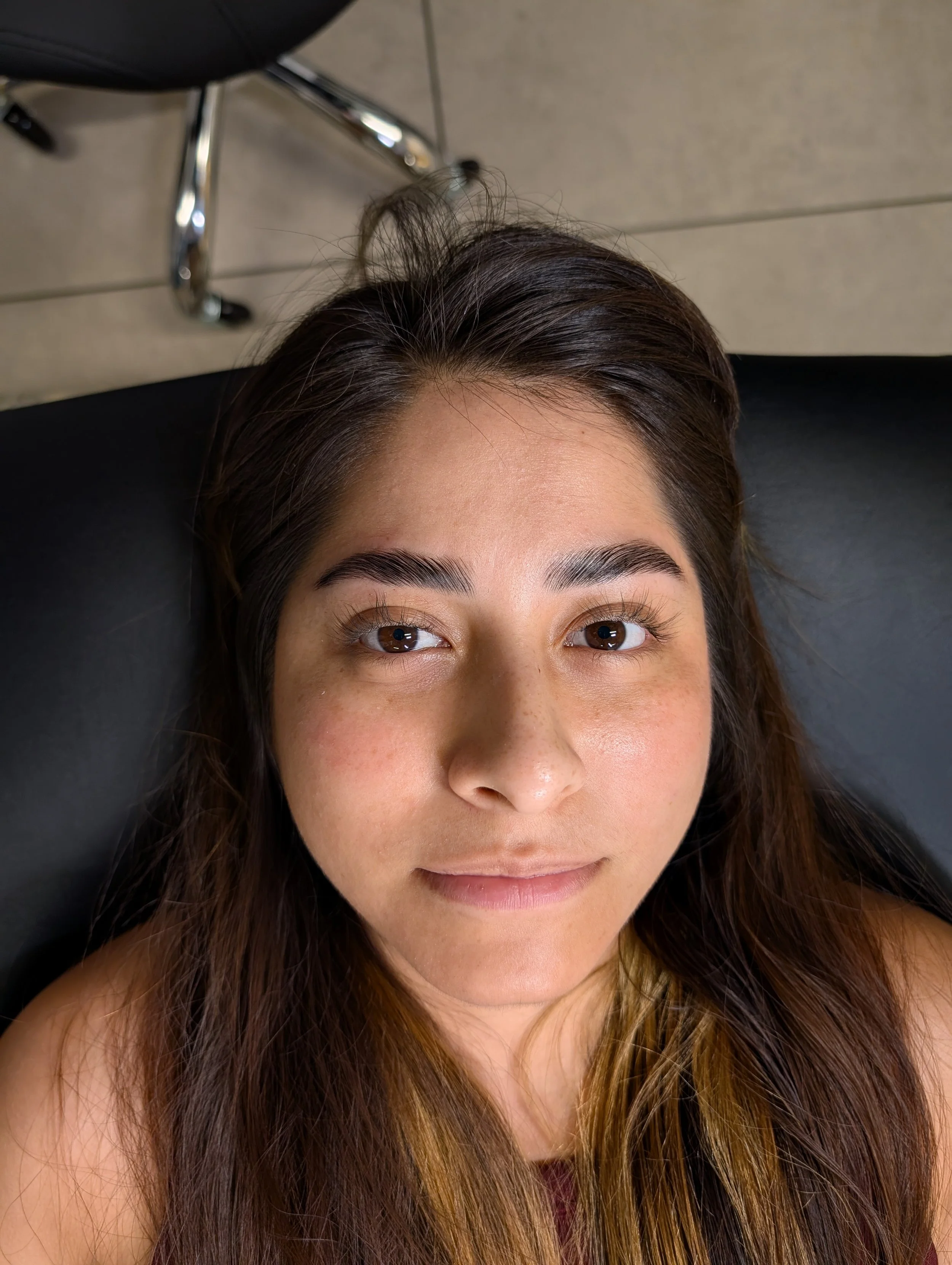 Close-up of a woman's face with brown eyes and long brown and blonde hair, looking up at the camera, with a gray floor and part of a black chair in the background. after lash lift and brow lamination