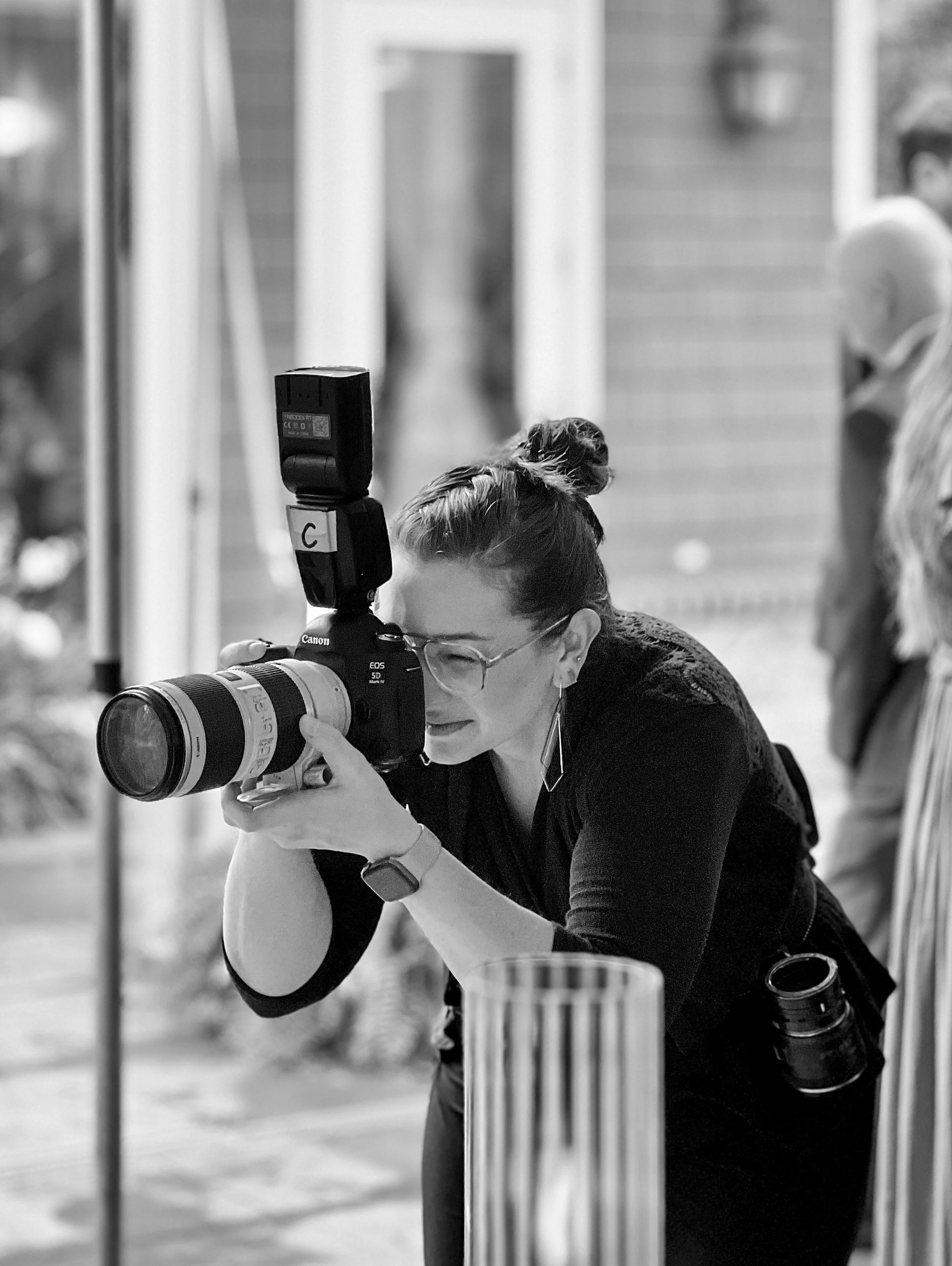 A woman with glasses and hair in a bun takes a photo with a professional camera in an outdoor setting, with people and a building in the background.
