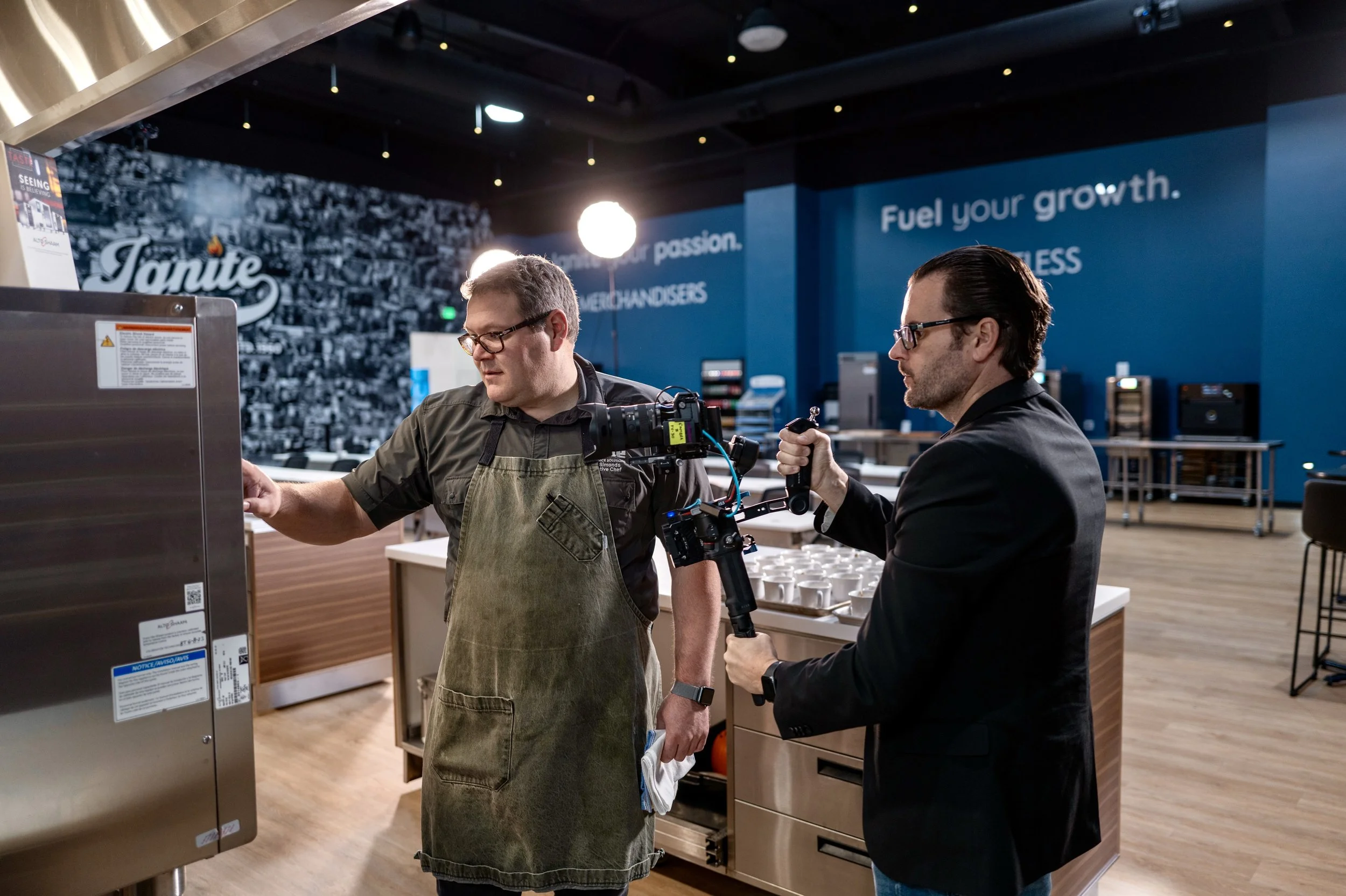 Two men are in a commercial kitchen. One is wearing an apron and is operating a commercial oven, while the other is holding a camera stabilizer and appears to be filming him. The background features a blue wall with the words 'Fuel your growth.'