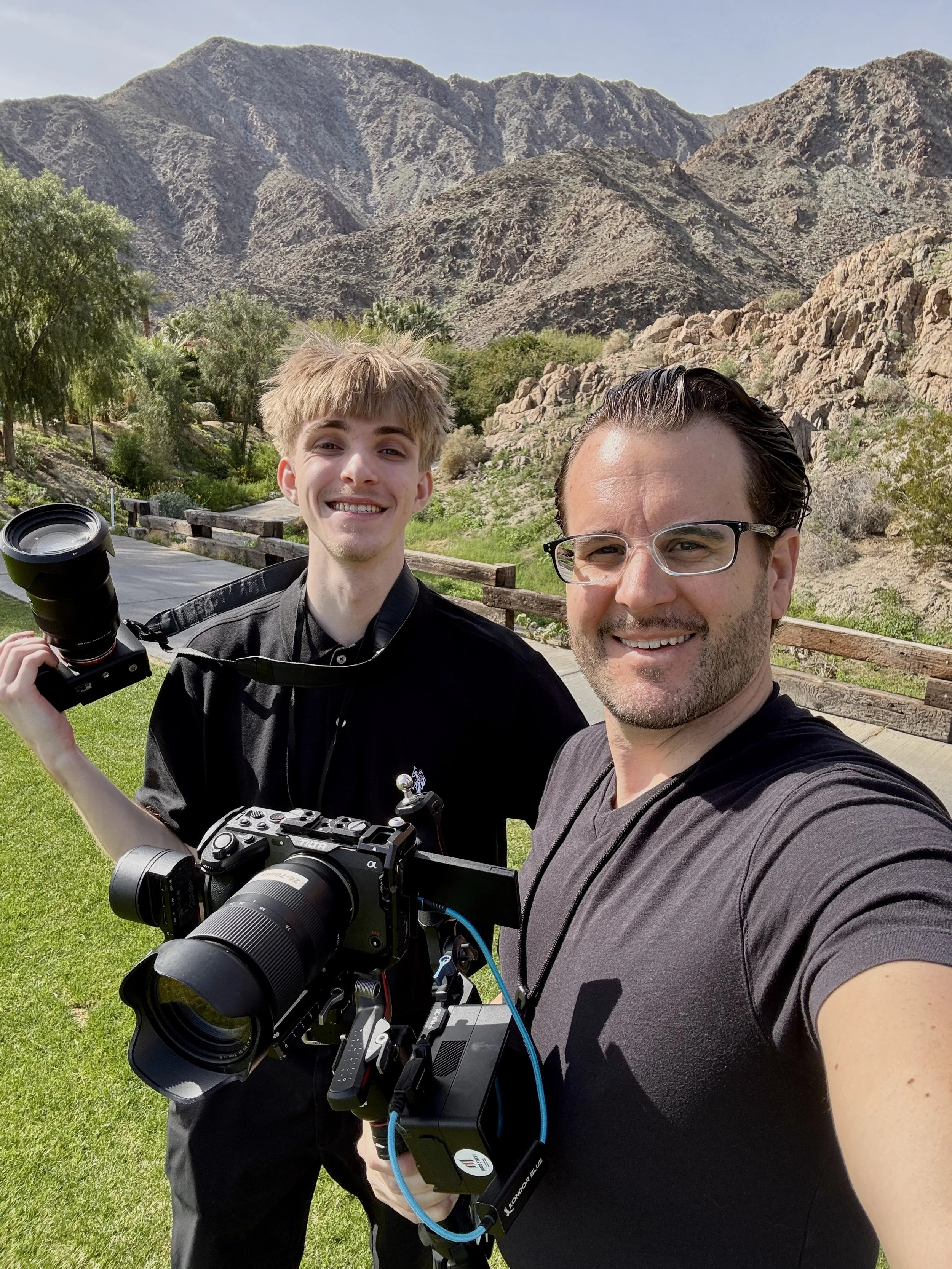 Two men taking a selfie outdoors in a mountainous desert landscape with green trees, rocky hills, and a clear sky.